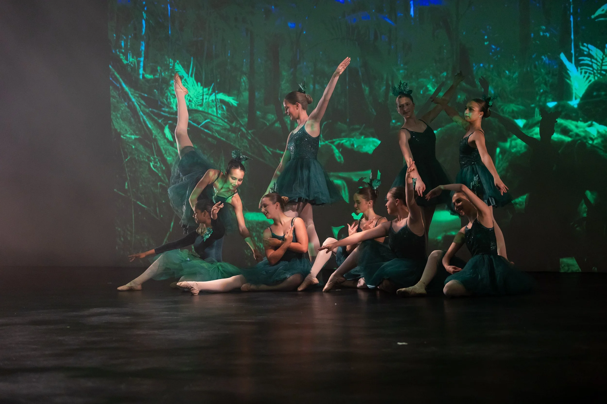 Group of young ballet dancers performing on stage with a forest-themed backdrop.