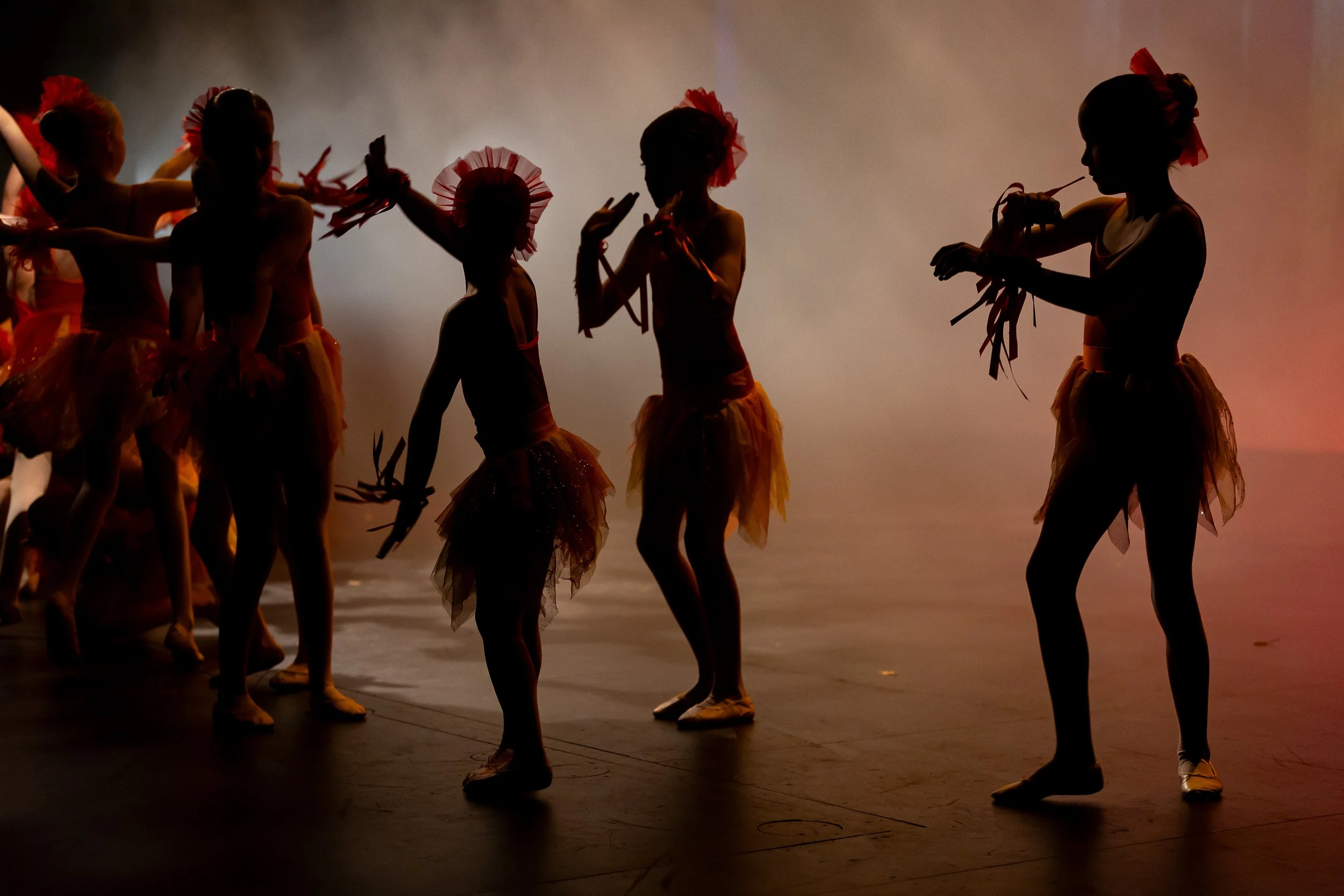 Silhouettes of young ballet dancers wearing tutus and headbands during a dance performance on stage with a dark background.