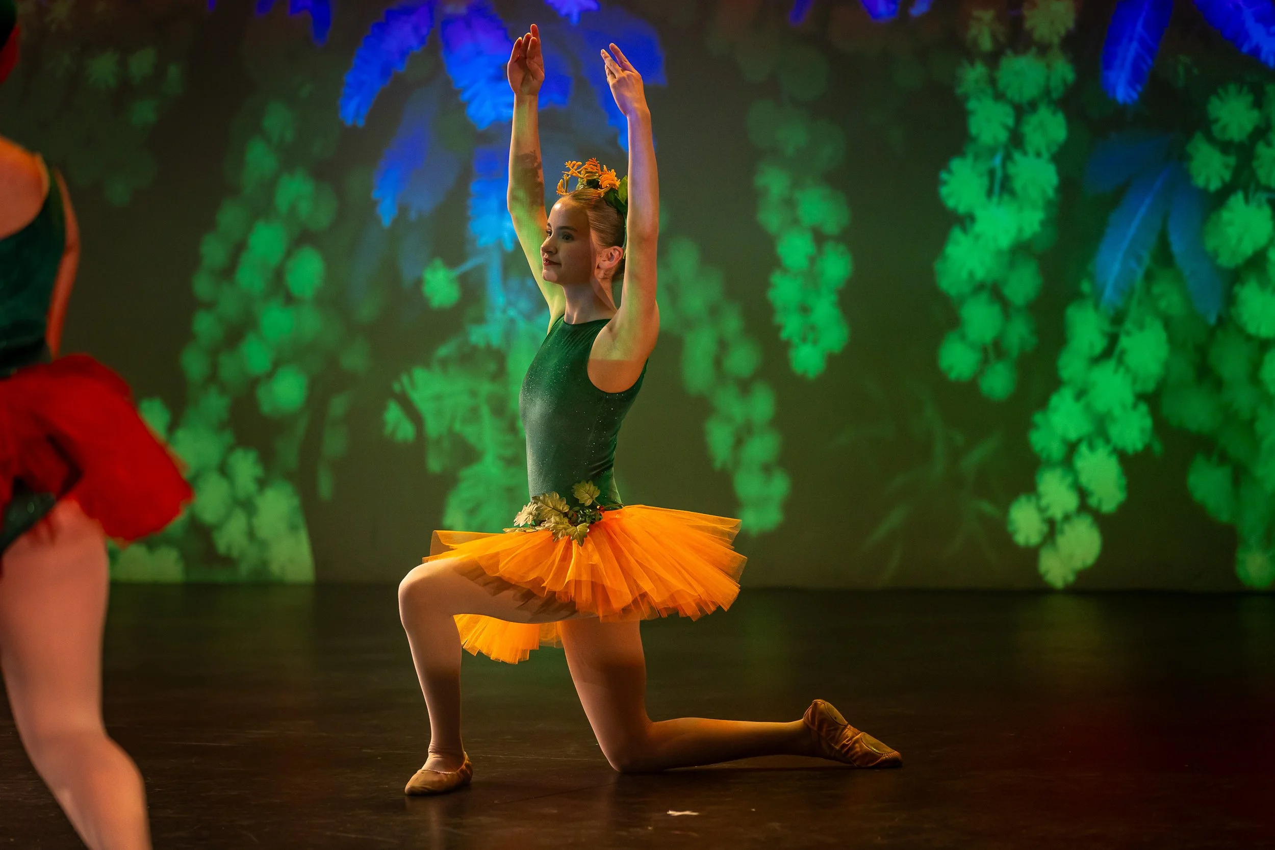 Young ballerina in a green and orange tutu performs on stage with a green and blue floral background.