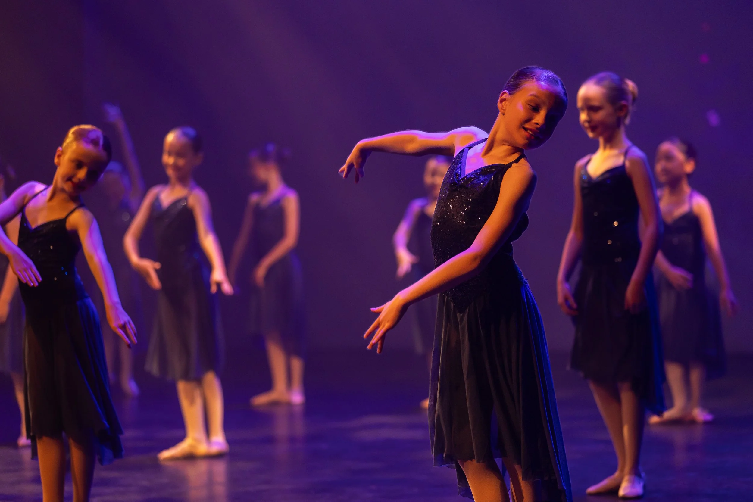 A group of young ballet dancers performing on stage, dressed in black costumes, under purple and blue stage lighting.