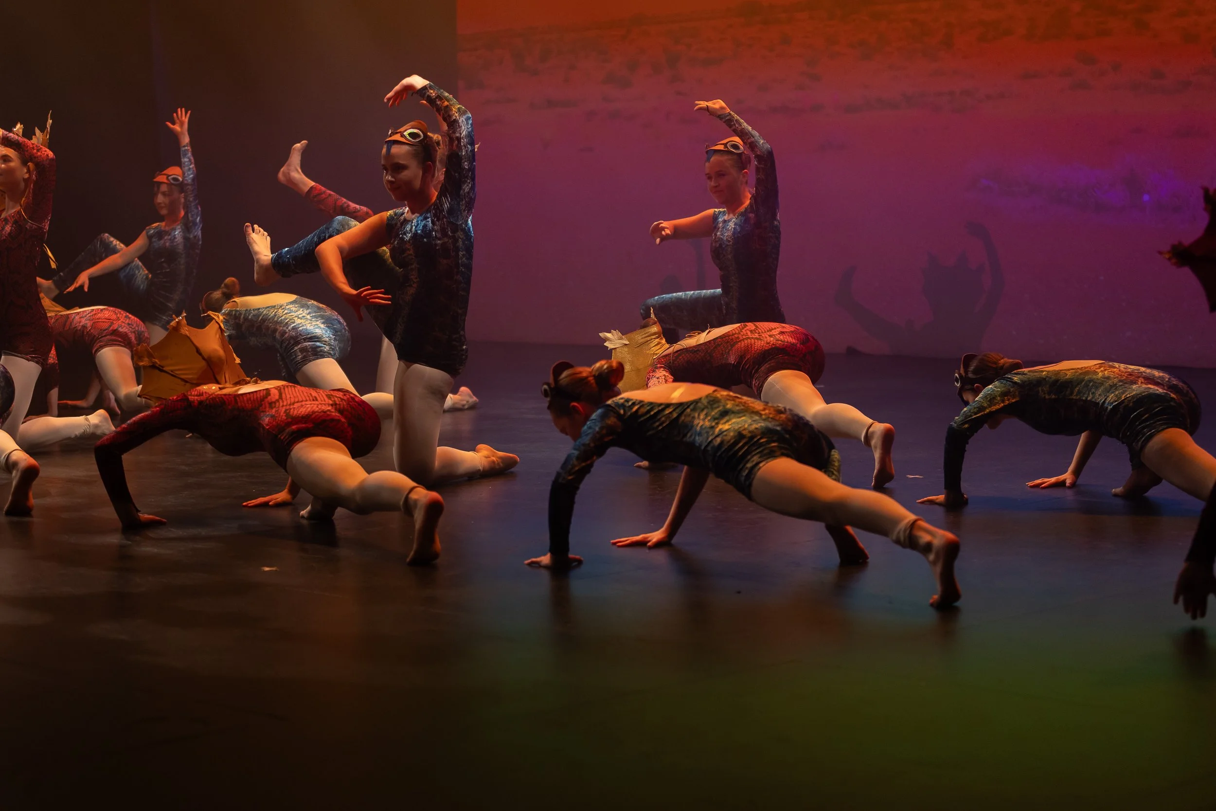 Group of young dancers in colorful costumes performing a ballet routine on stage with a dark background and colorful lighting.