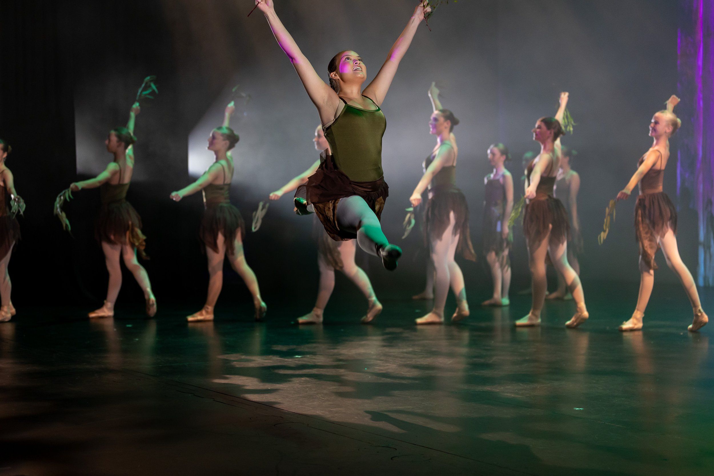Group of dancers performing on stage with one dancer mid-air in the foreground, all dressed in black costumes.