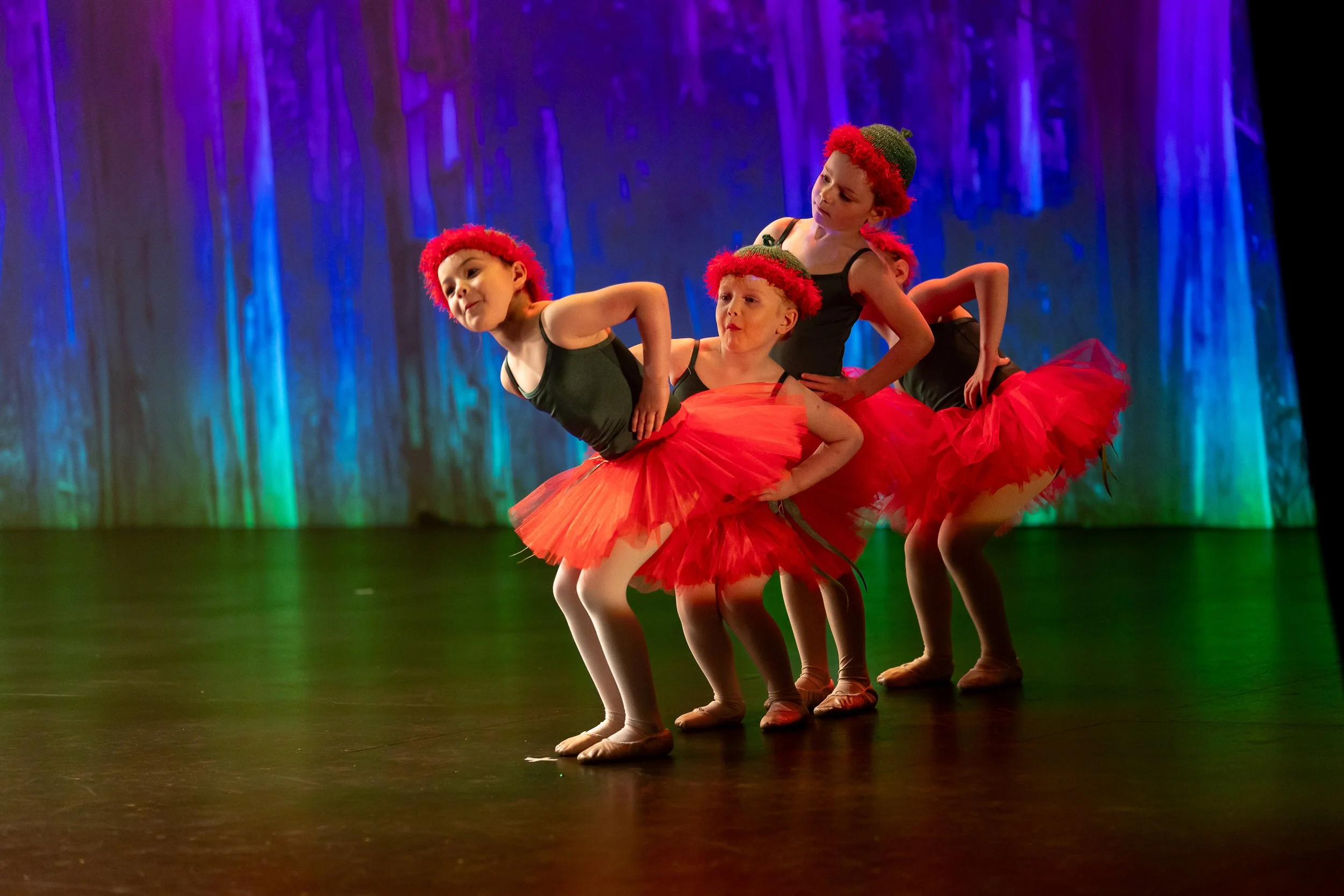 Four young girls dressed as strawberries perform a ballet dance on stage with a colorful forest backdrop.