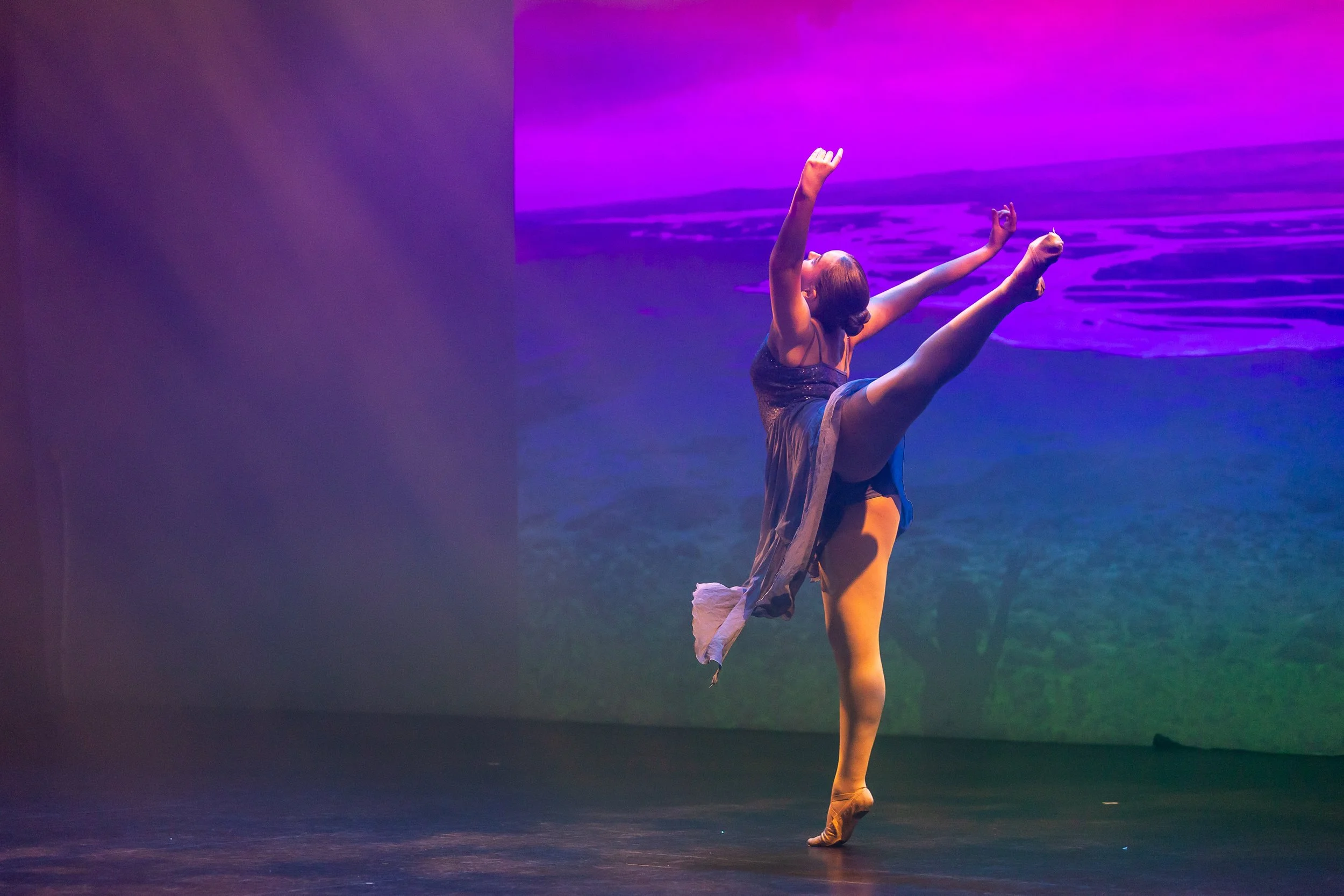 Ballet dancer performing on stage with a colorful scenic background, balancing on one leg with arms raised and extended.