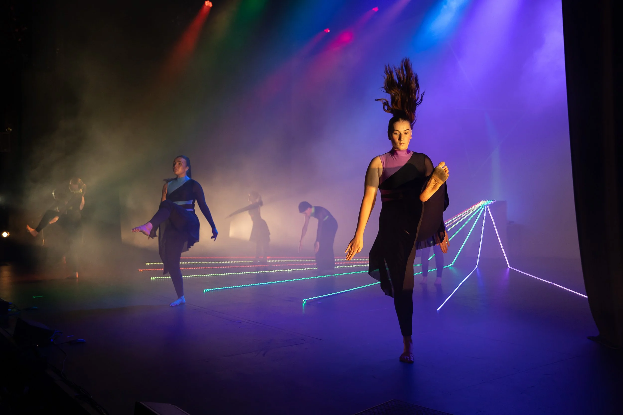 Dancer with flowing hair performs on stage with colorful lighting and fog, wearing a purple and black costume with one arm covered in a brown glove, while other dancers are in the background.