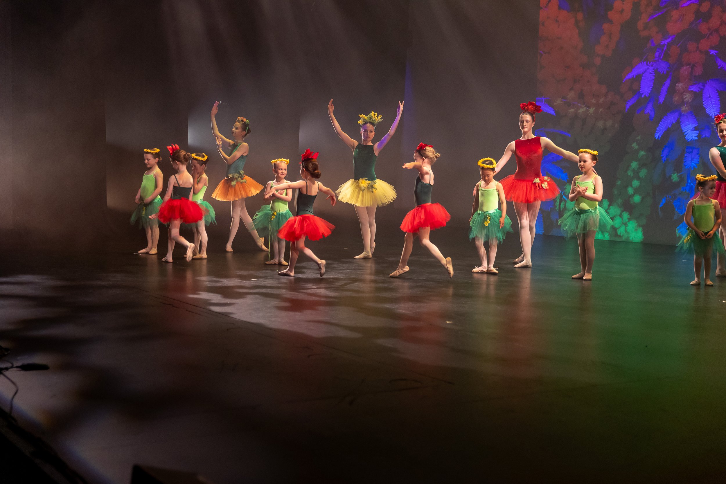 Group of young ballet dancers performing on stage, wearing colorful tutus and floral headbands, with bright stage lighting and background projection.