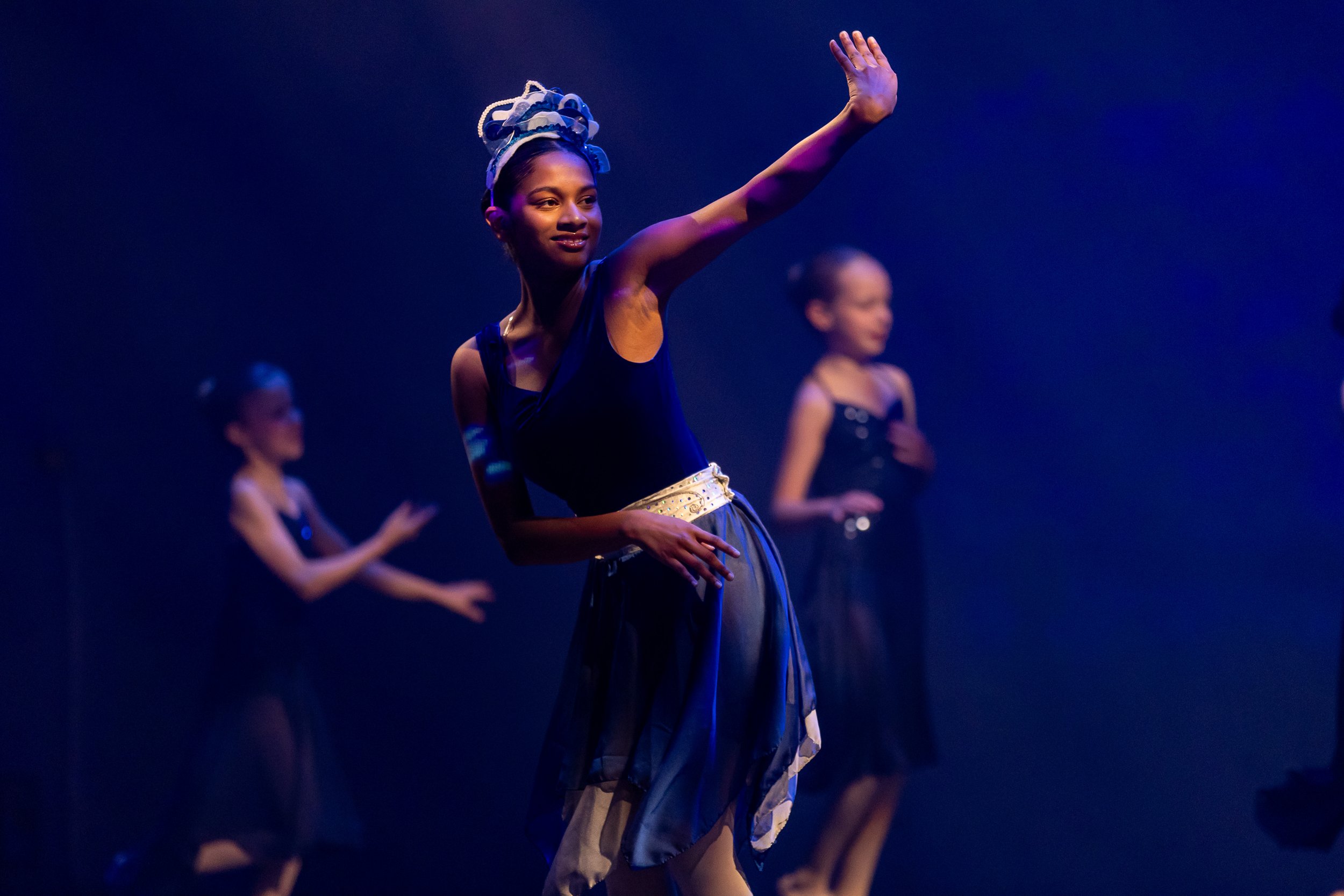 Dancers performing on stage wearing black and beige costumes, with a lead dancer in the foreground raising her hand, illuminated by stage lighting.