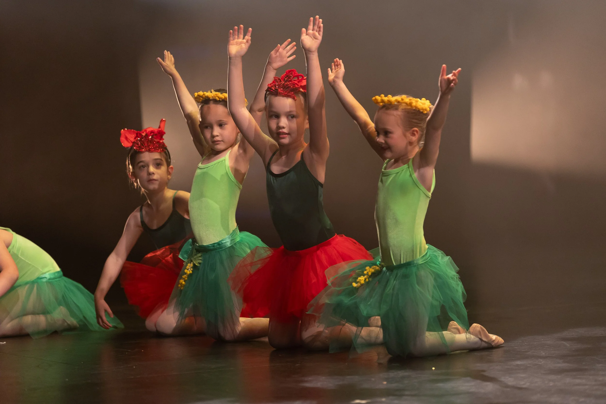 Group of young girls dressed in colorful ballet costumes performing on stage, kneeling with arms raised.