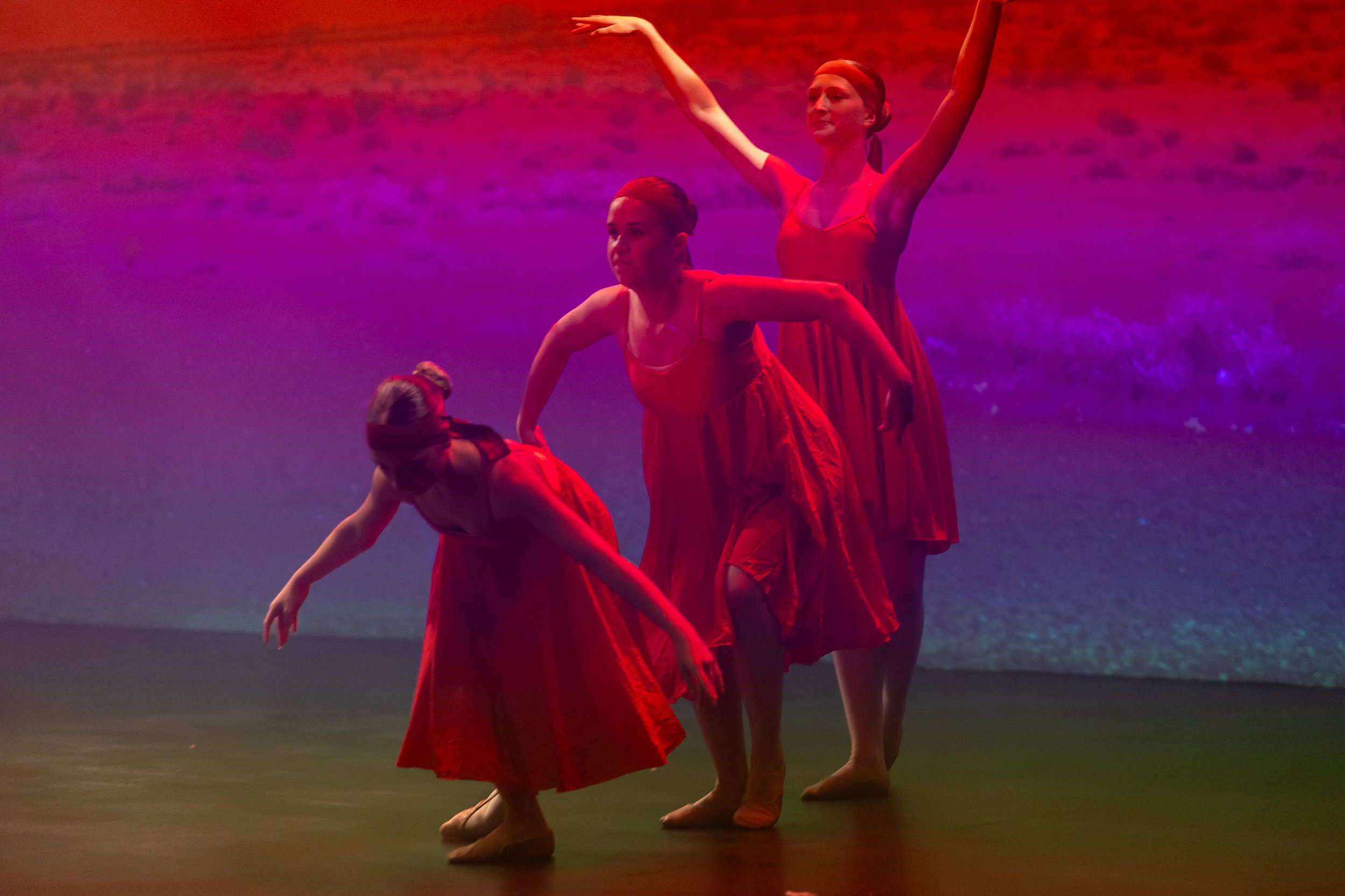 Three young women in red dresses performing a dance on stage with a beach scene projected in the background.