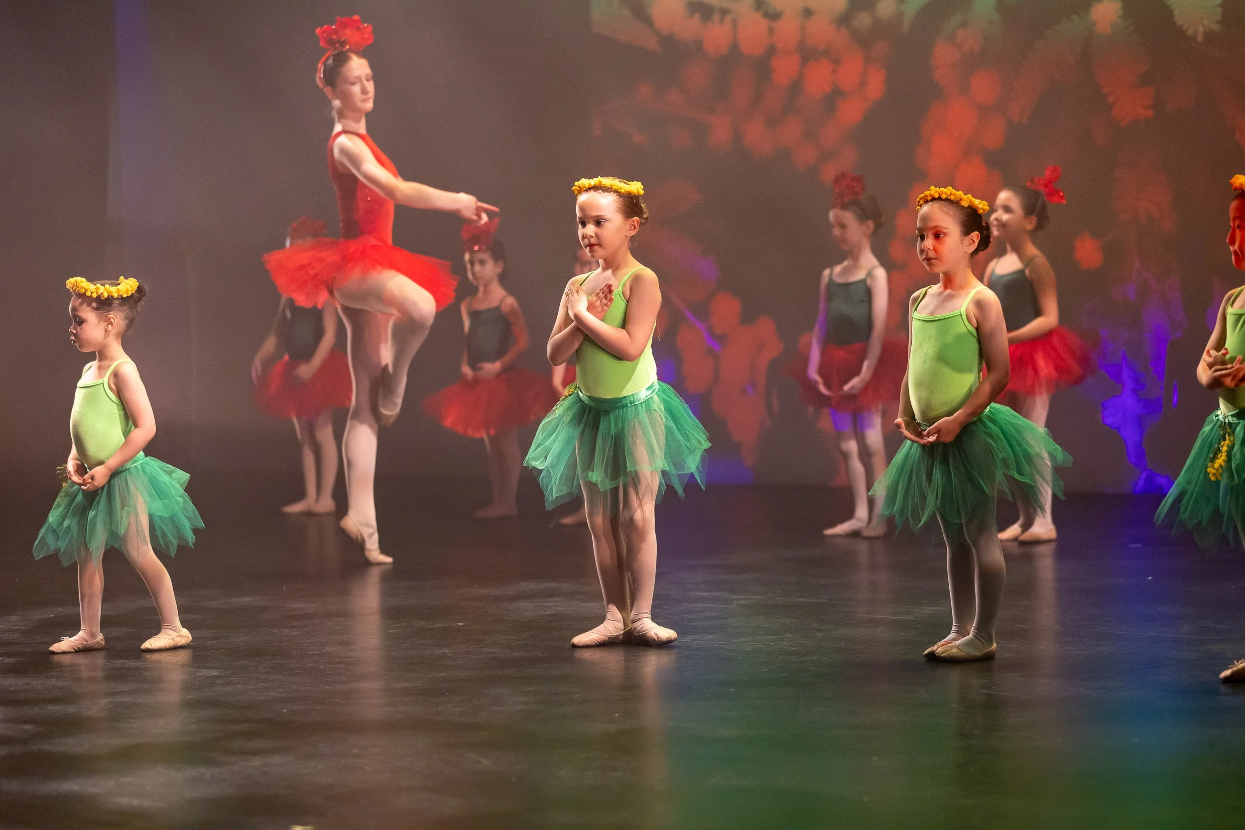 Young ballet dancers dressed in green tutus and yellow flower crowns perform on stage, with a backdrop of red foliage, during a dance recital. An instructor in a red costume demonstrates a ballet pose among the children.