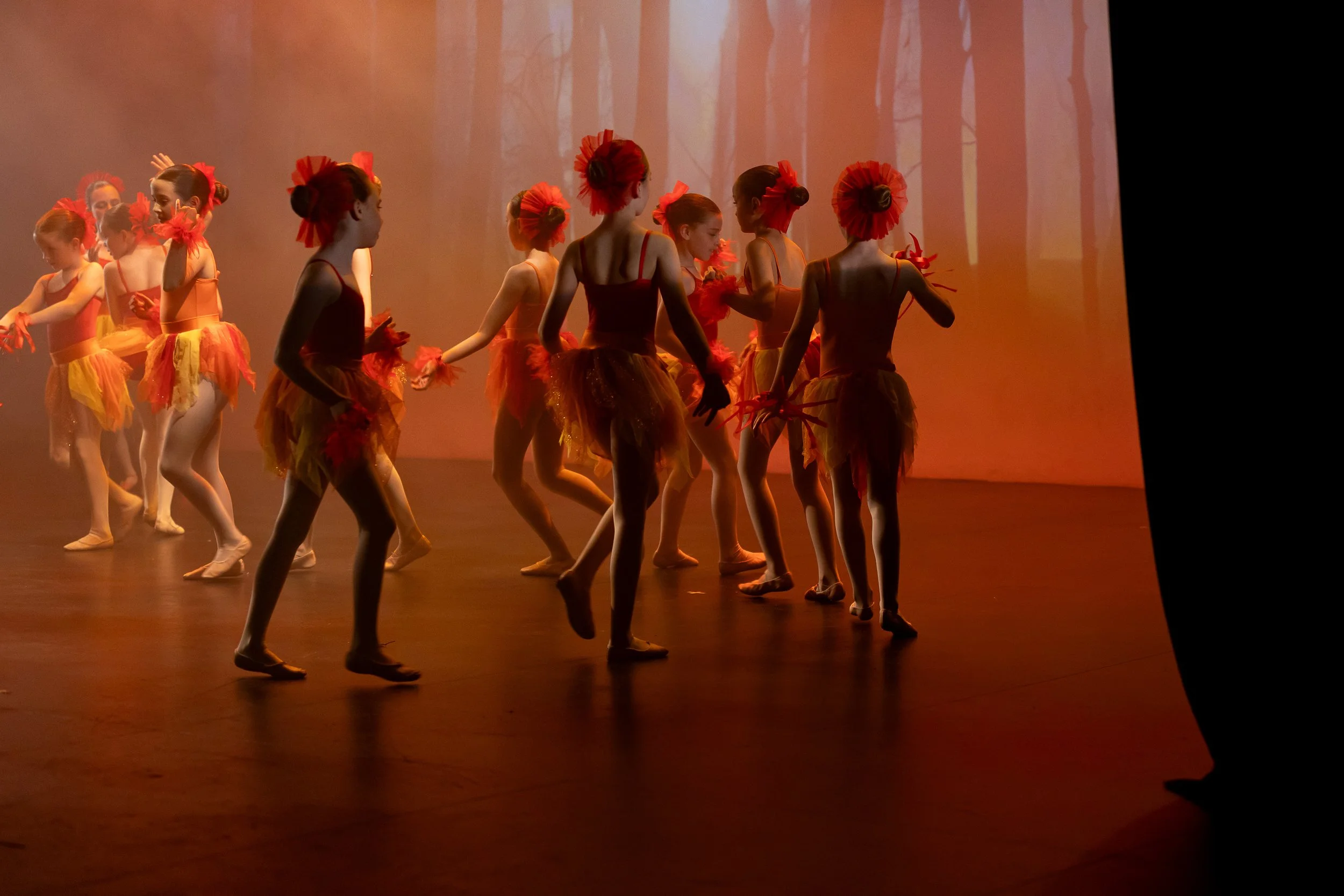 Group of young girls in colorful costumes performing a dance on stage with a forest backdrop projection.