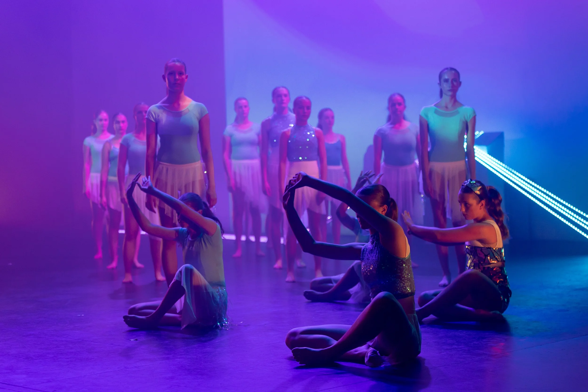 Young ballet dancers practicing on stage under colorful purple and blue lighting.