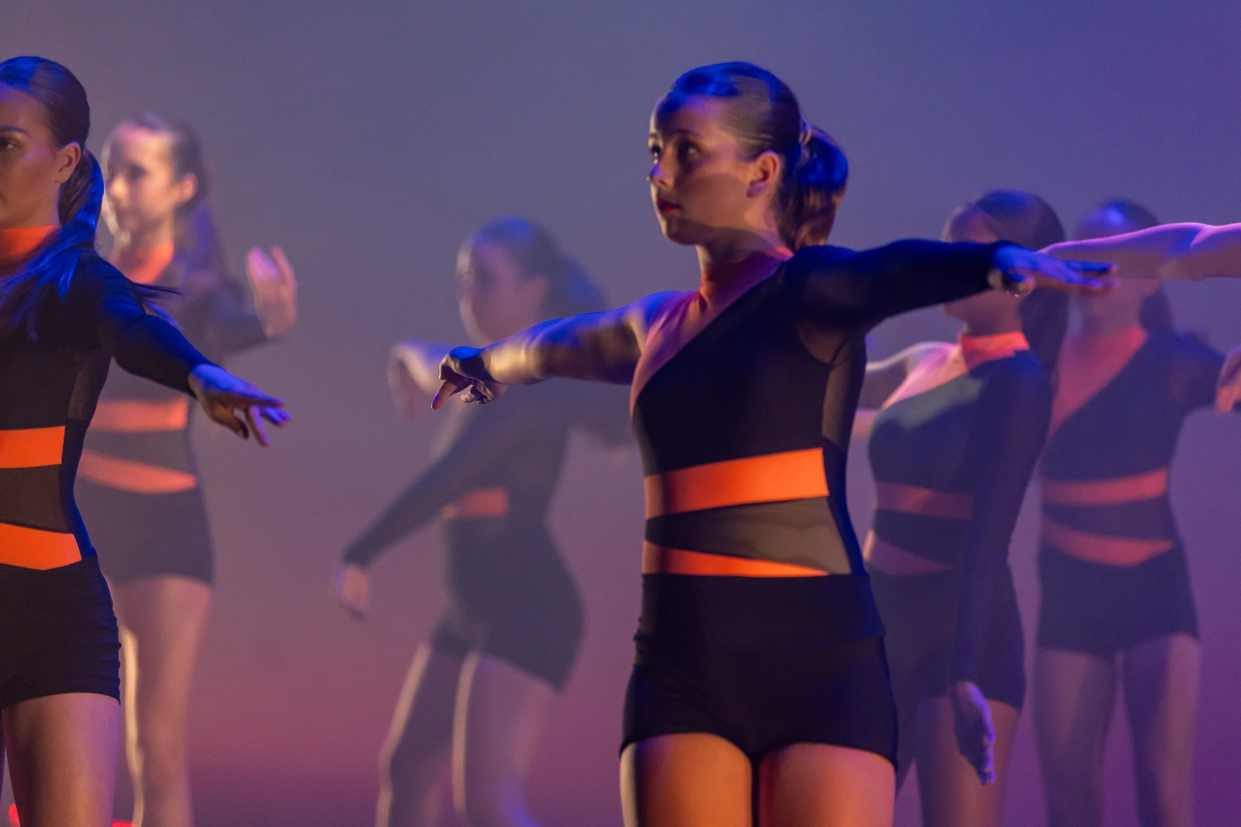 A group of dancers in black and orange costumes performing on stage with arms extended.