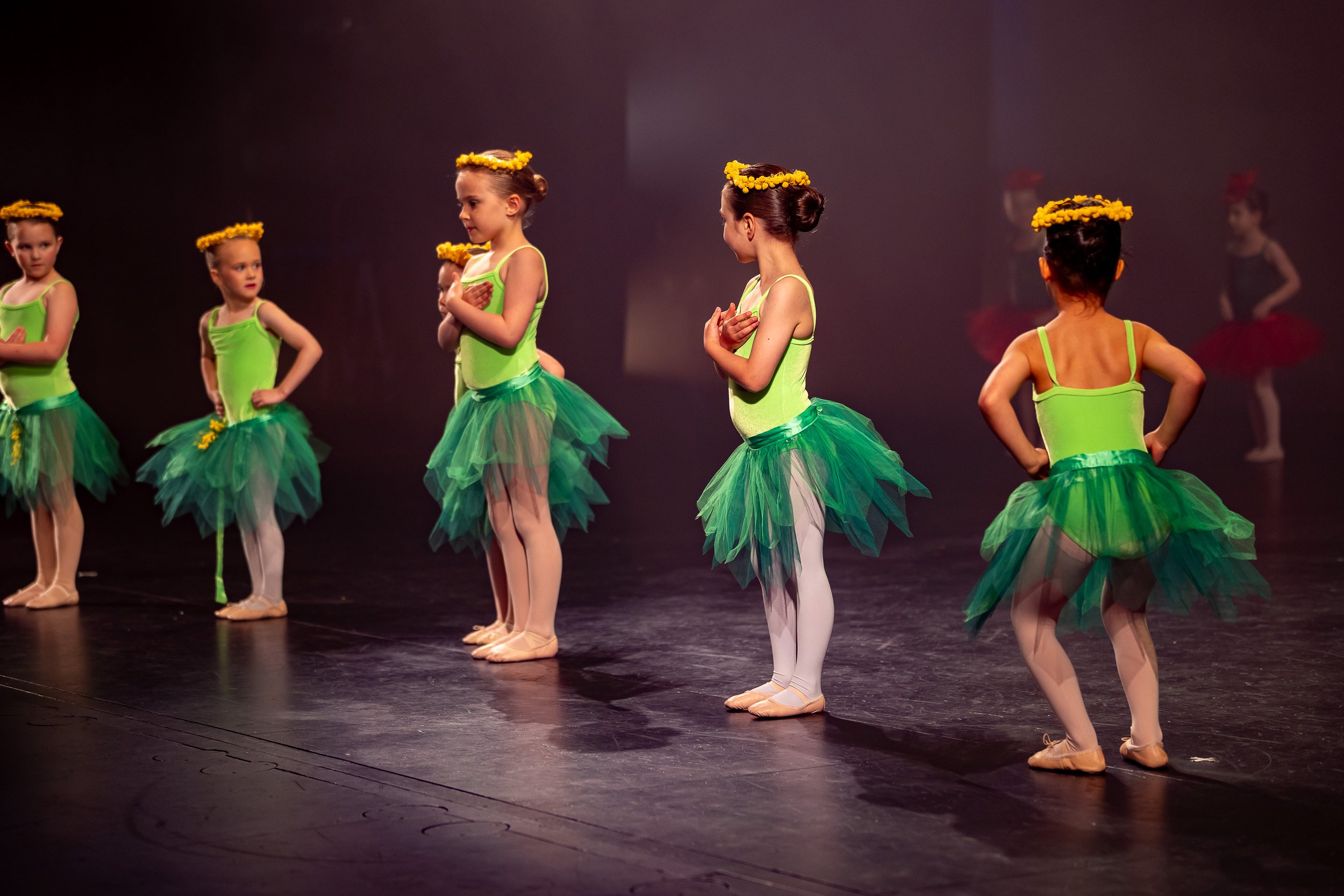 Young girls in green ballet costumes with yellow flower crowns perform on stage with a dark background.