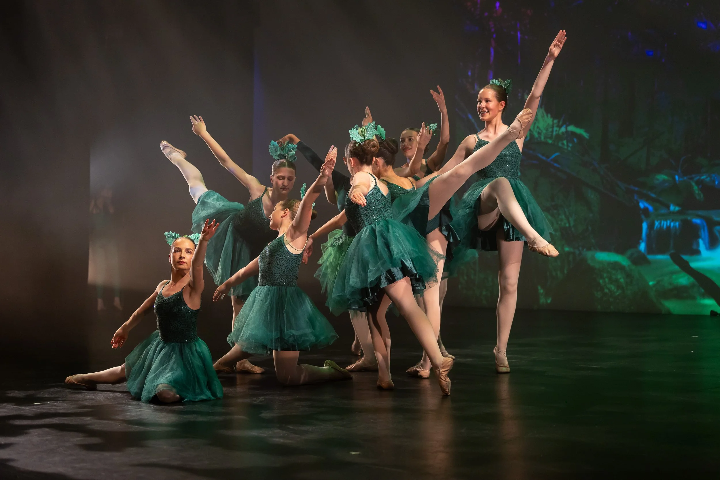 Group of ballet dancers performing on stage in green costumes with a nature-themed backdrop.