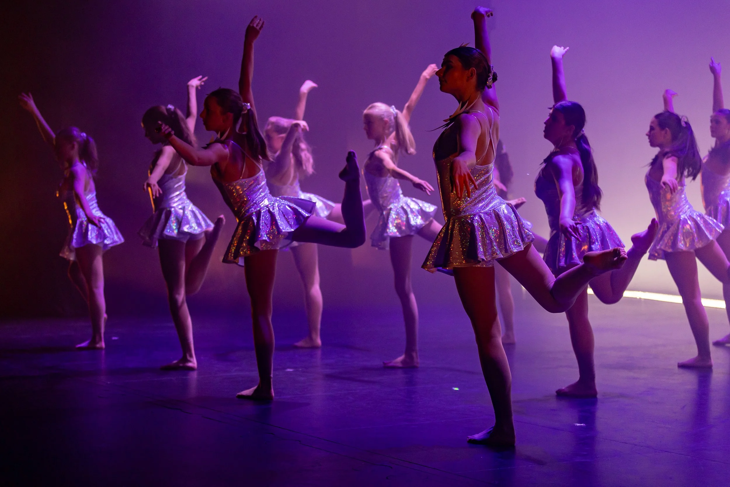Group of young girls performing a dance on stage, wearing shiny silver dresses and striking various dance poses under purple stage lighting.