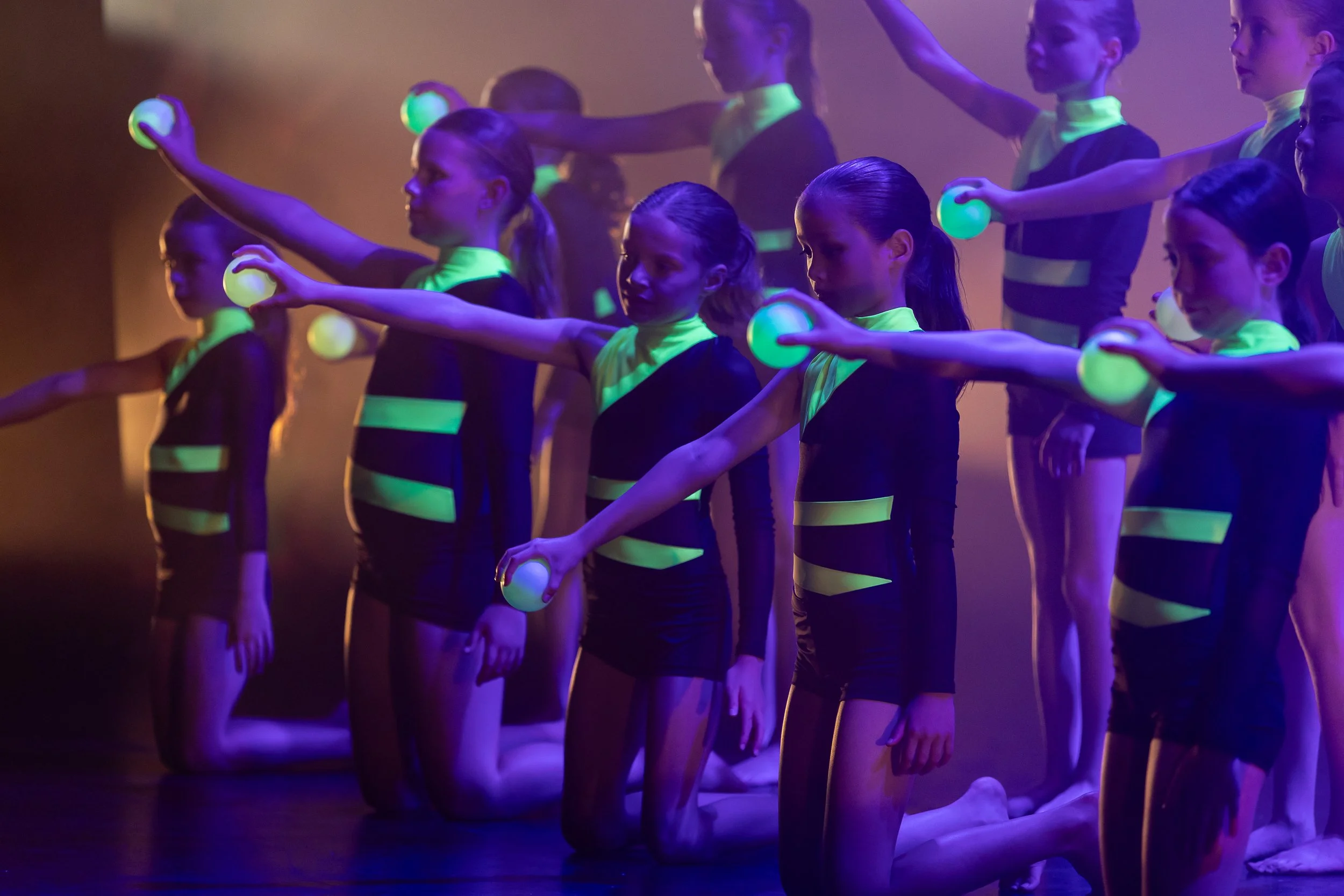A group of young girls dressed in black with neon green accents performing a dance routine under blacklight, holding glowing balls, with some kneeling and others standing.