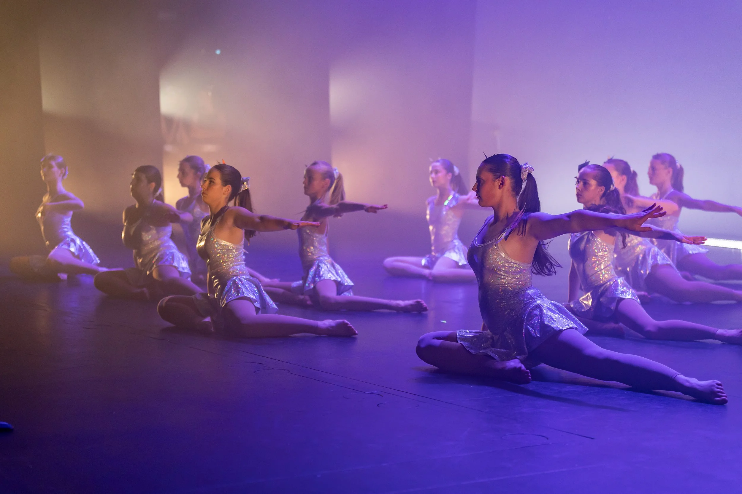 Dancer girls in shiny silver costumes performing a routine on stage with purple lighting.