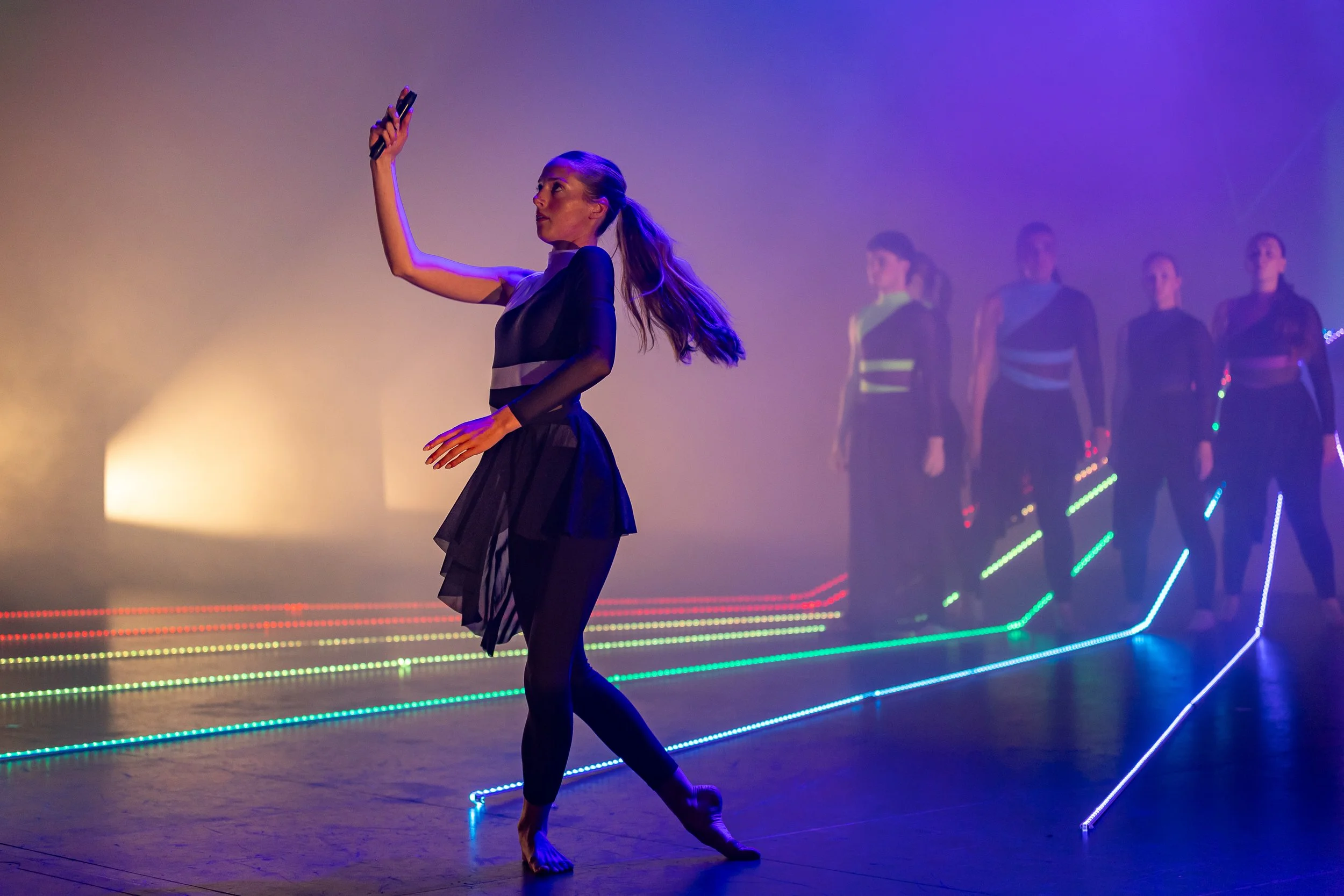 A woman in a black dress takes a selfie in a performance space with neon lights and four women in similar outfits standing behind her.