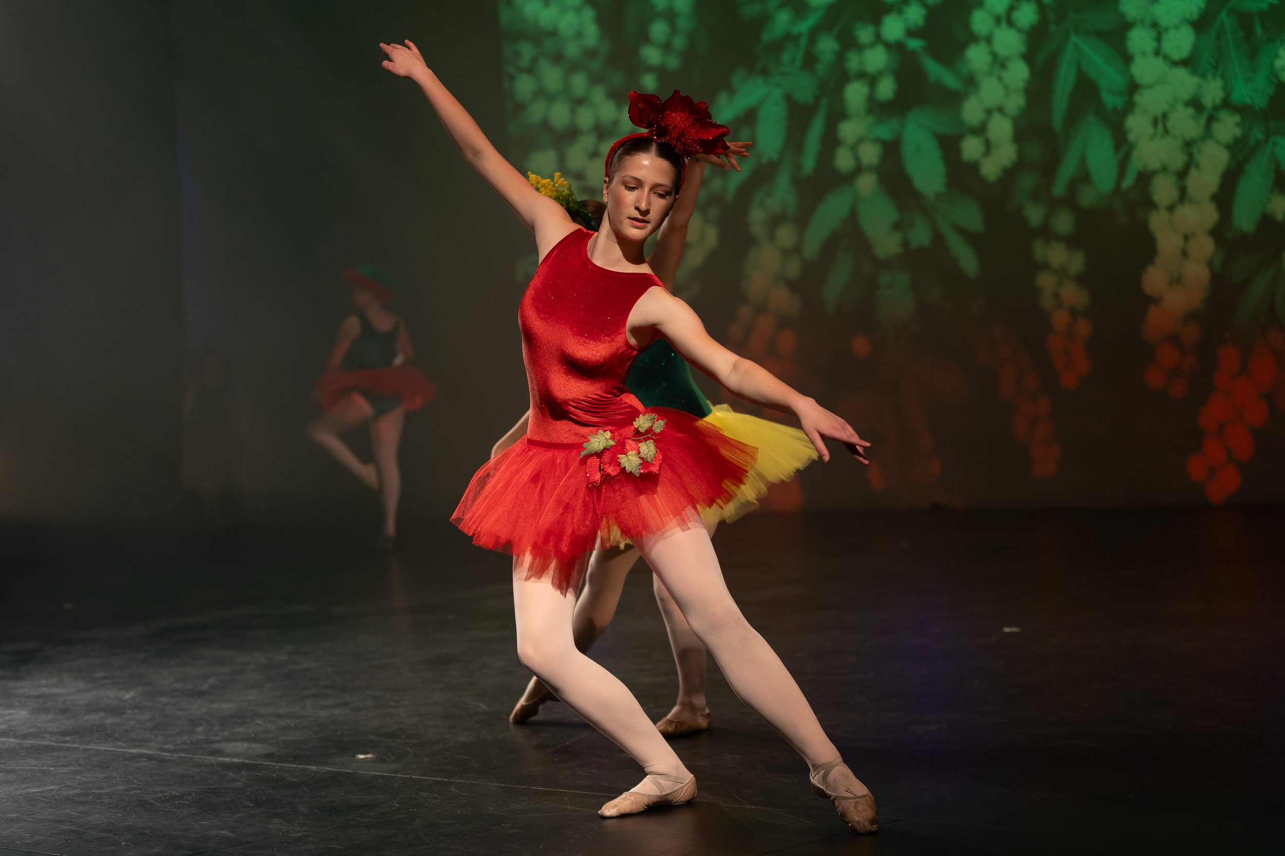 Ballet dancer in a red and yellow tutu with floral decorations performing on stage with a green and red background.