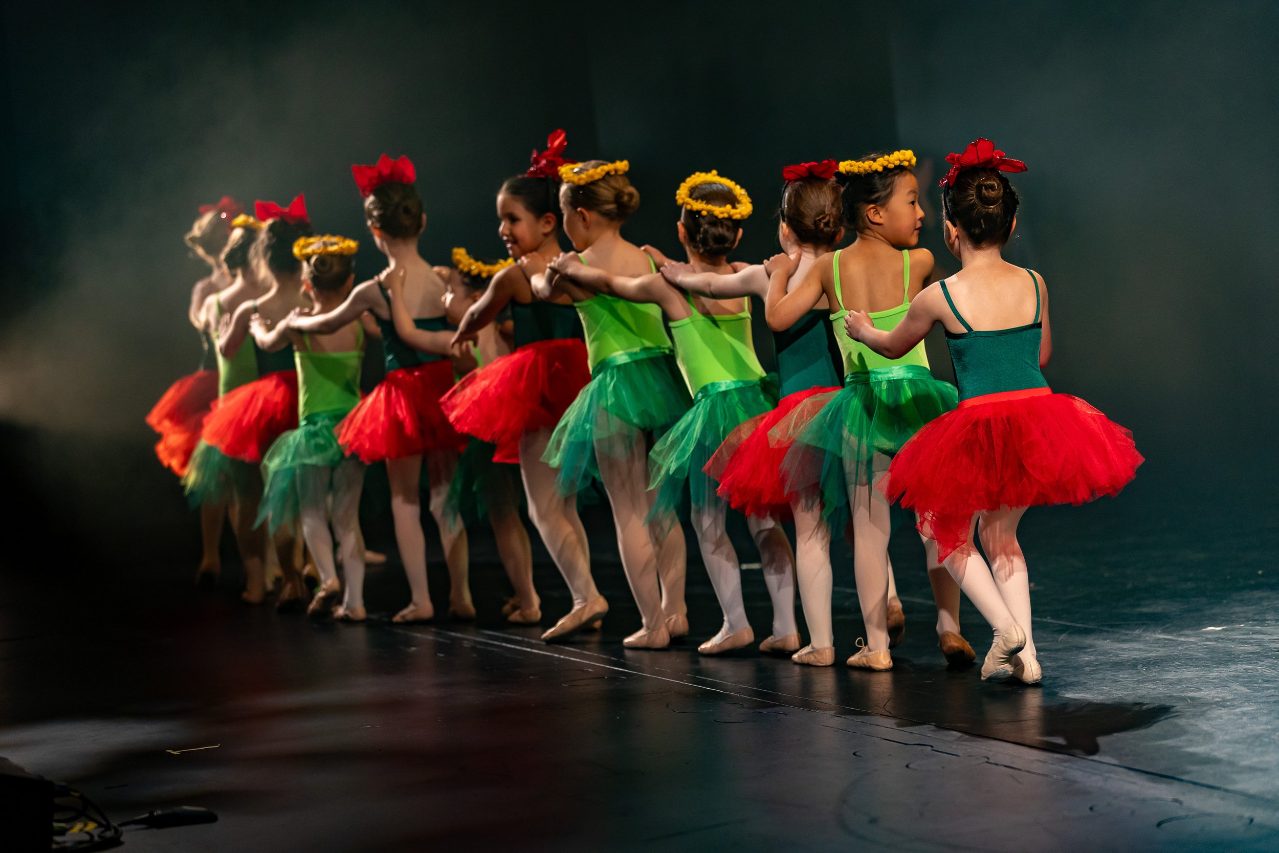 Group of young girls in colorful ballet costumes standing in a line on stage, holding shoulders, with dark background and stage lighting.