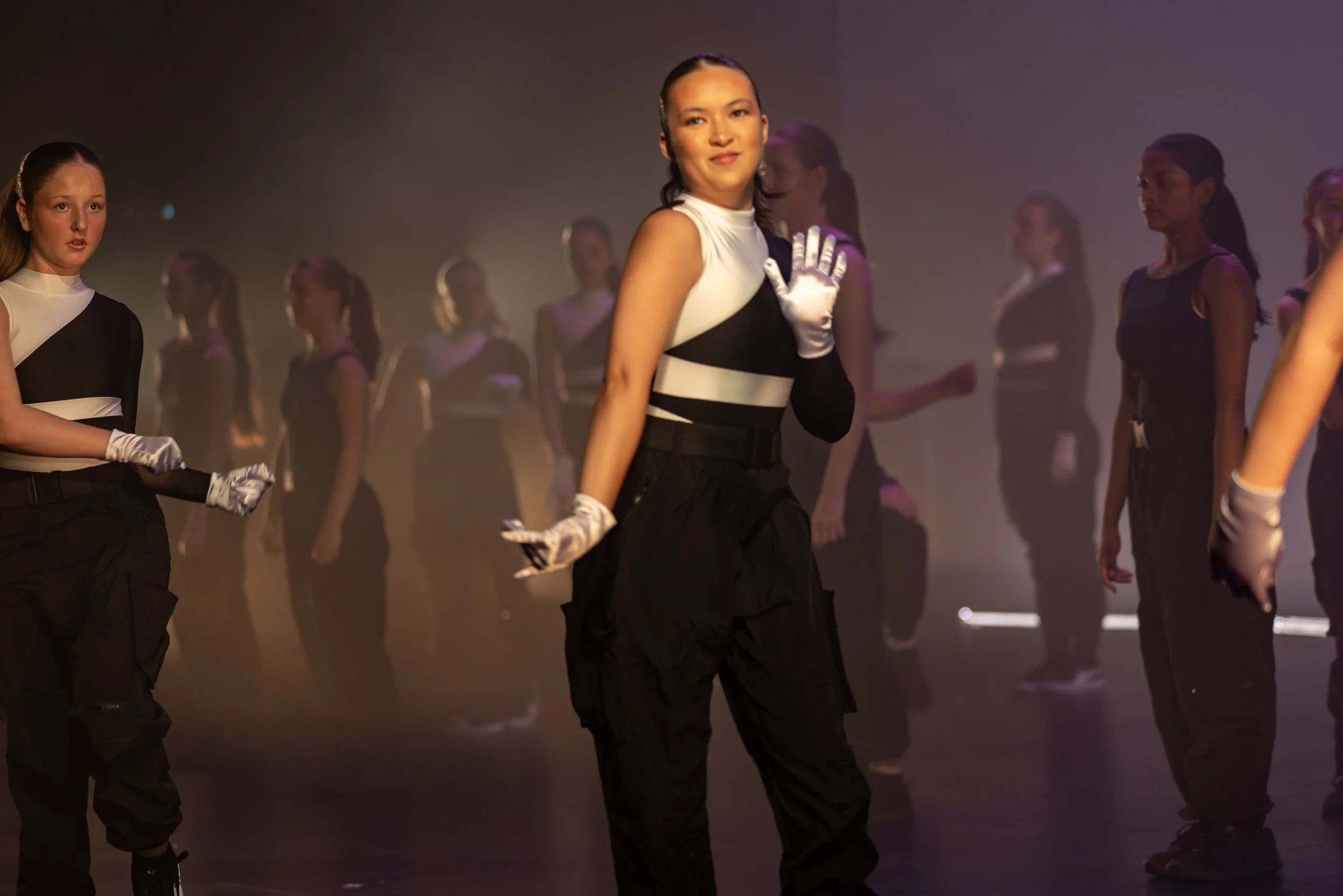 A group of young women in coordinated black and white outfits and white gloves, standing on stage in a dance or performance setting with atmospheric lighting.