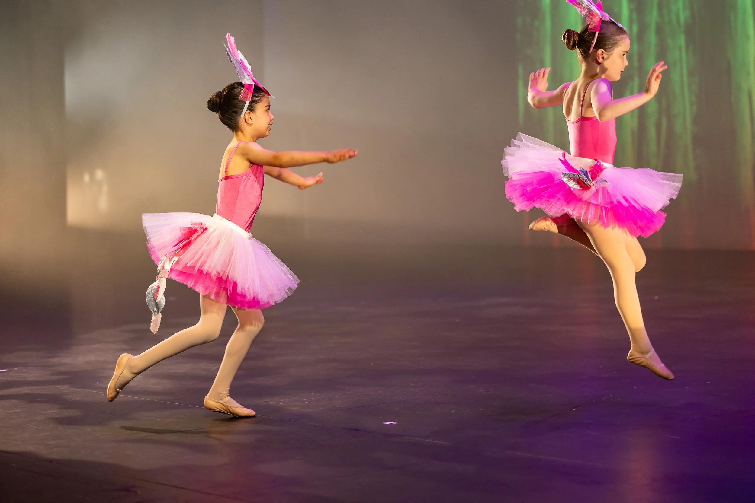 Two young girls dressed as ballerinas performing on stage with pink tutus, pink leotards, and pink headpieces with feathers, mid-jump during a dance routine.