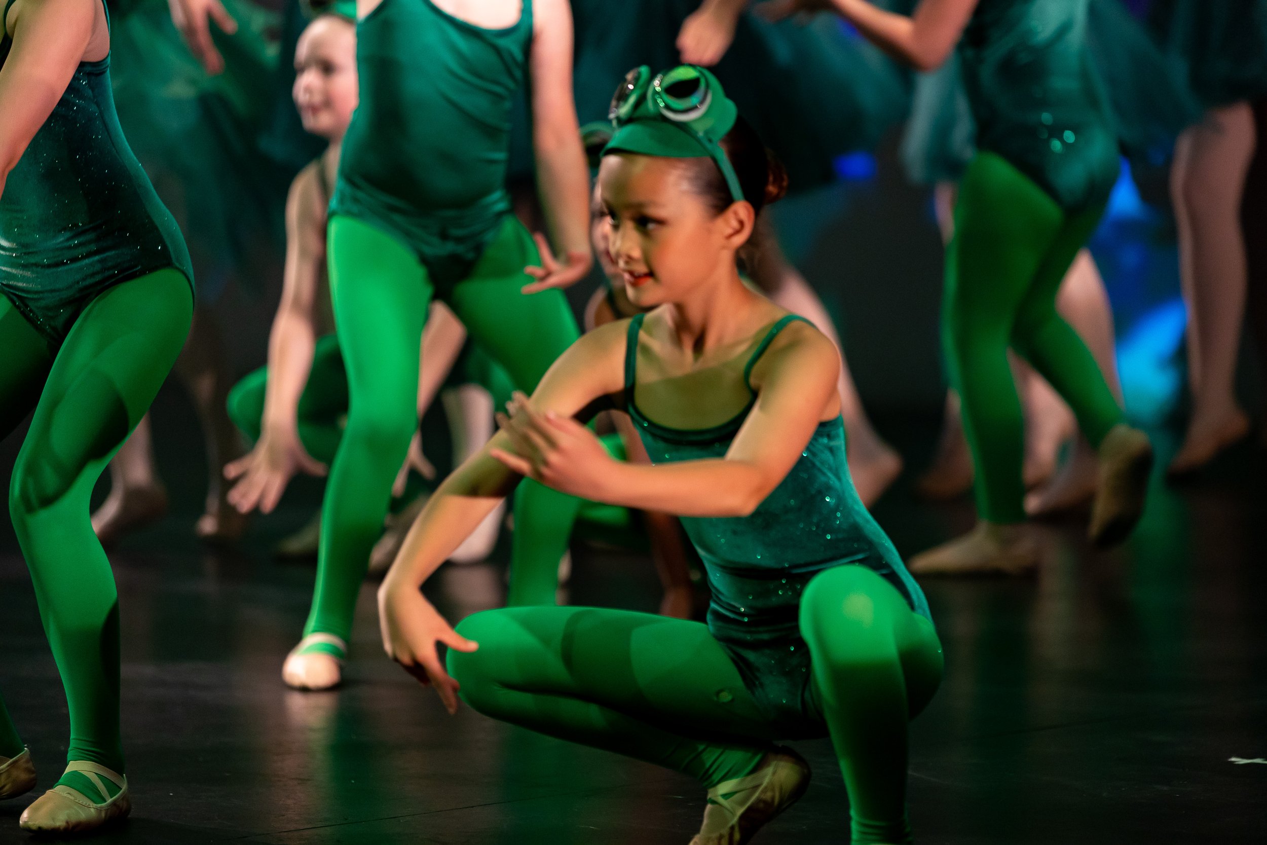 A young girl in a green dance costume with sparkles, wearing green tights and dance shoes, and a headband with goggles, performing a dance move on stage with other dancers in matching costumes.