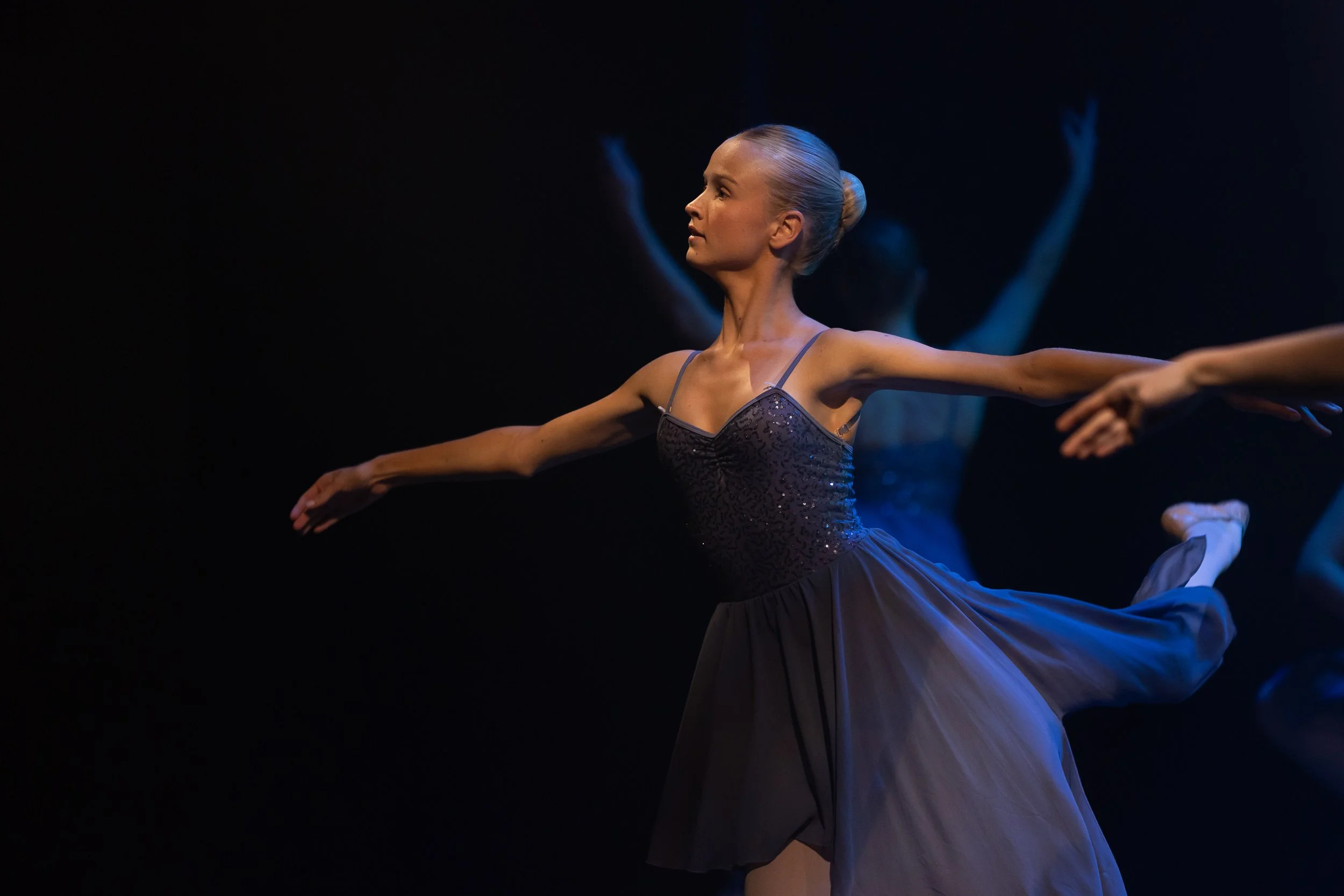Ballet dancer performing on stage in a dark setting, wearing a black dress and balancing on one leg with arms extended.