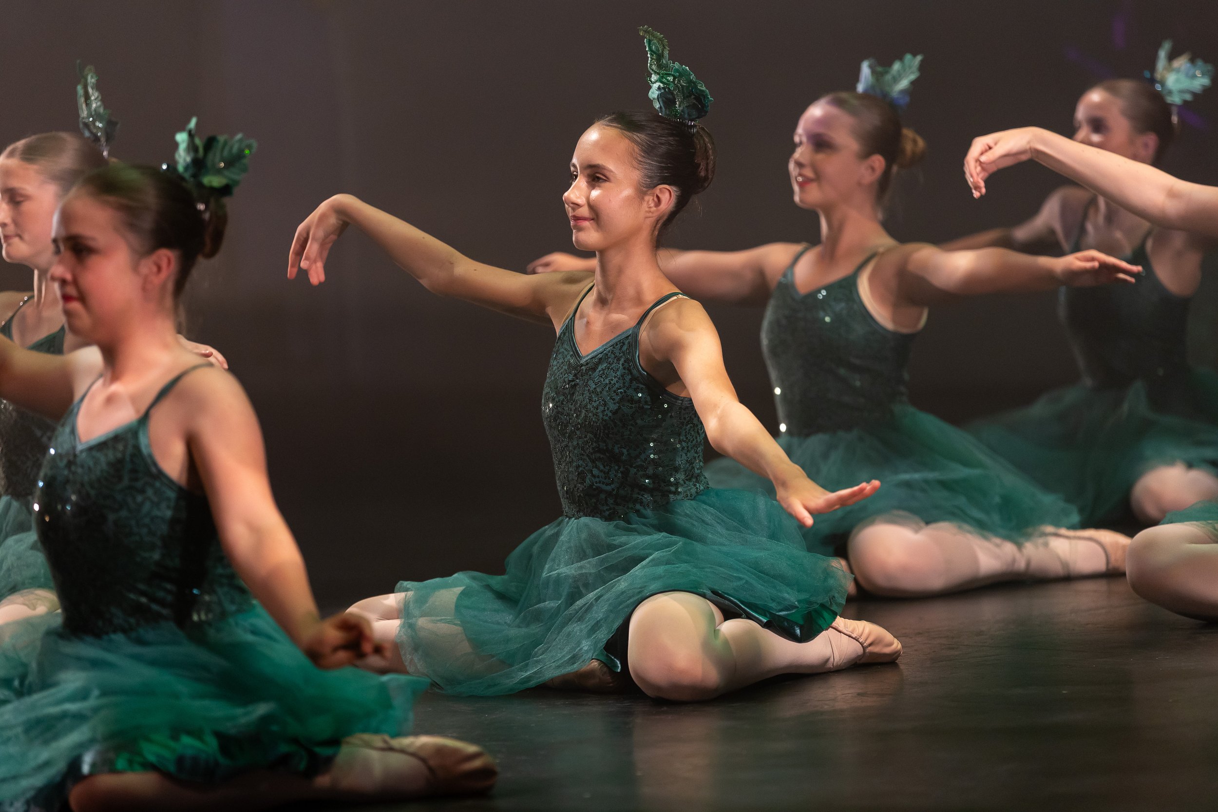 A group of young ballet dancers in green costumes practicing on stage, sitting in a kneeling position with one arm extended and the other bent, performing a dance routine.