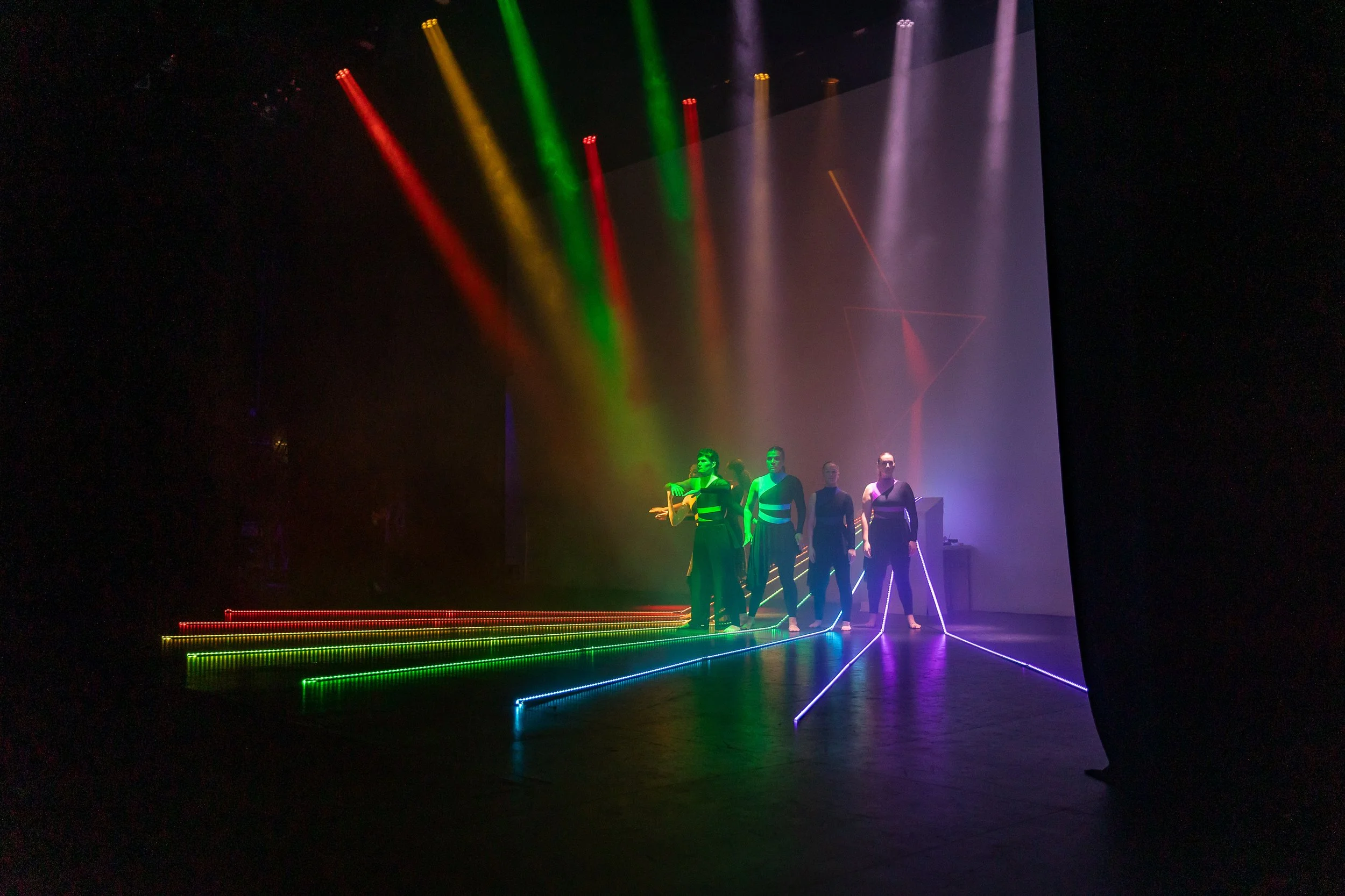 Four performers dressed in black with illuminated accents stand on a stage illuminated by colorful lights and neon lines, with a geometric projection in the background.