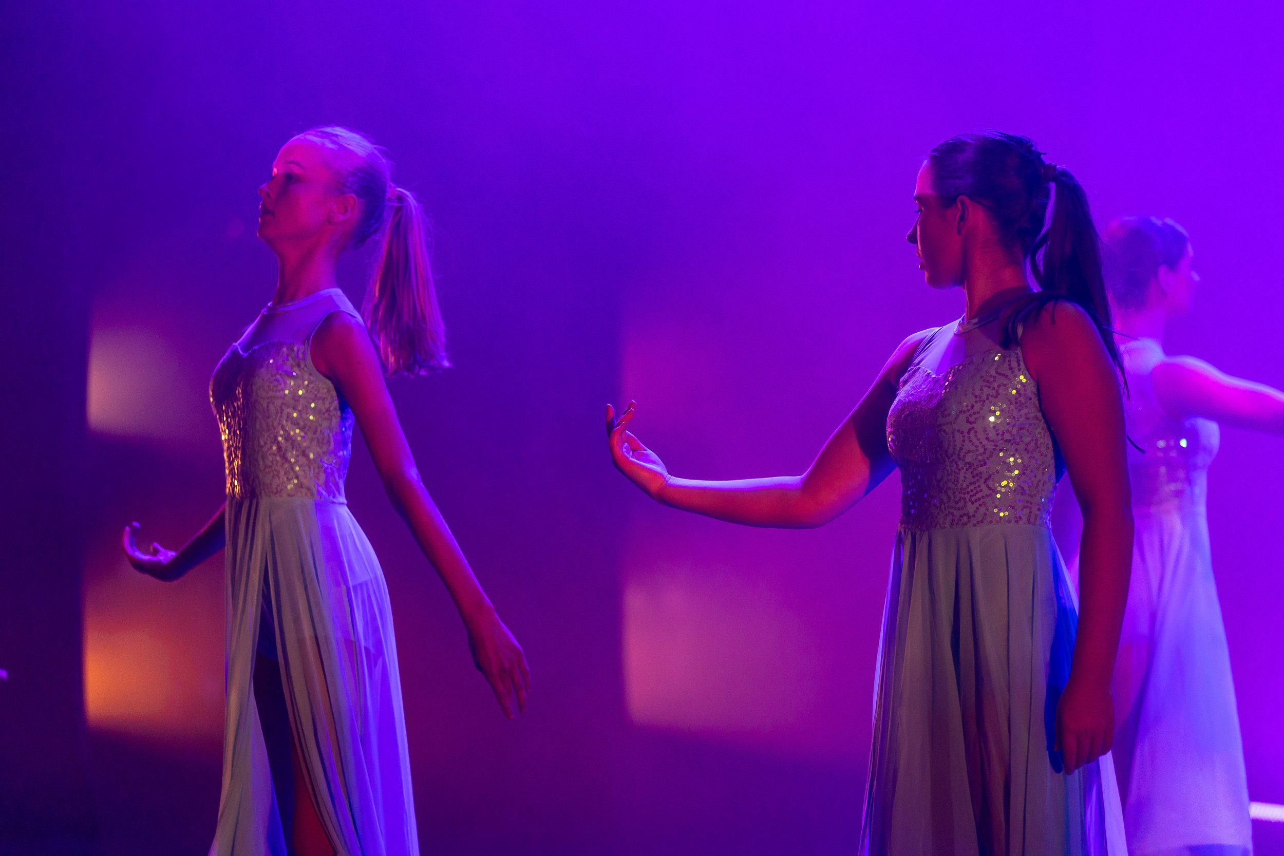 Three women in elegant dresses performing a dance or pose on stage under purple lighting.