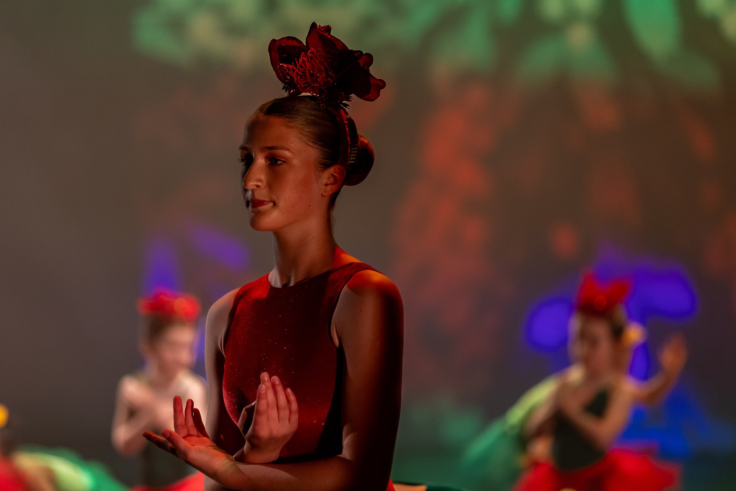 Young female ballet dancer in red costume during a performance, with others in similar attire in the background.