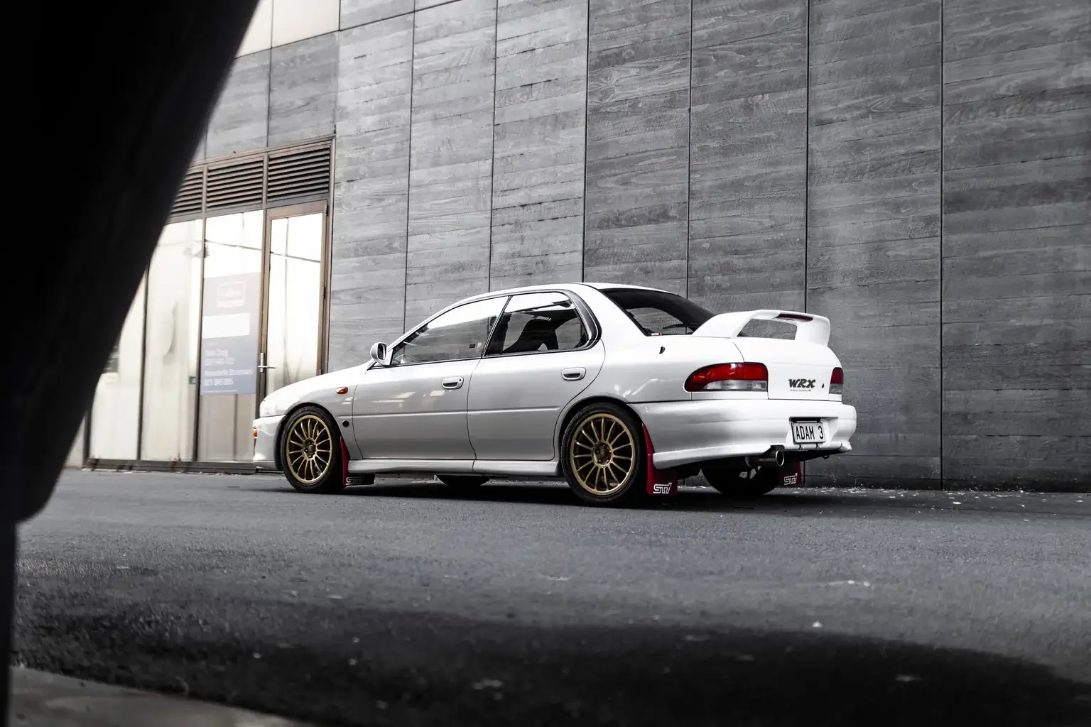 artistic shot looking through building pillar in industrial zone showing rear side shot of white gc8 subaru impreza WRX STi version 4 1998 JDM vehicle for export sale New Zealand