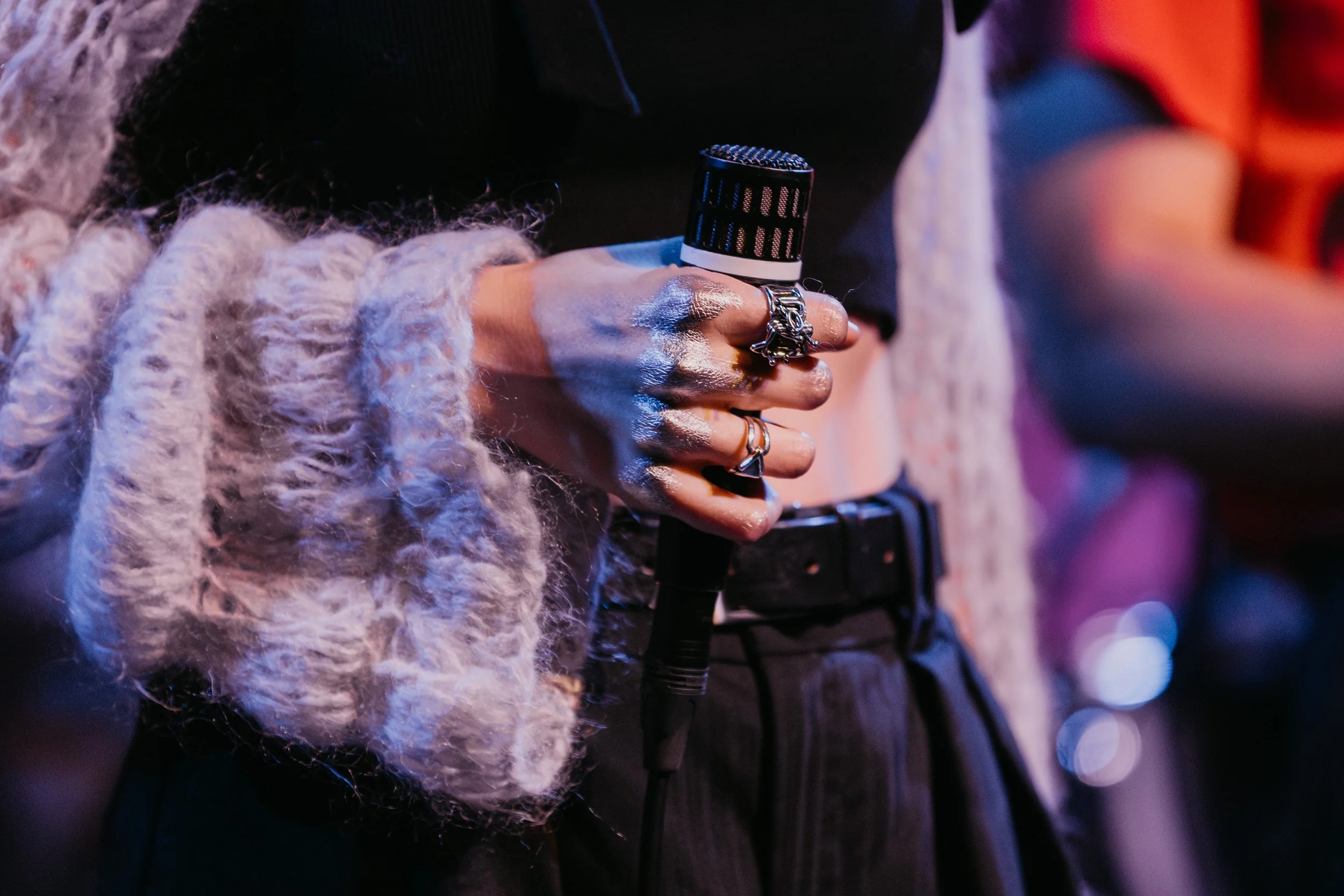 A person holding a microphone with multiple rings on their fingers, wearing a chunky knit sweater and dark pants.
