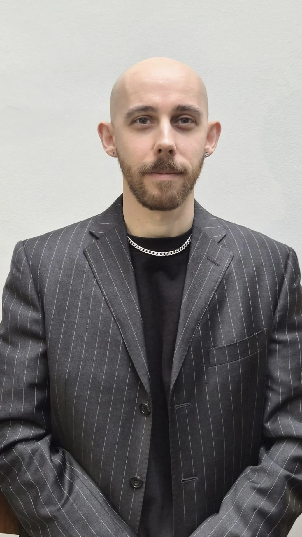 A man with a beard and mustache, bald head, wearing a dark pinstripe blazer, black shirt, silver chain necklace, and small earrings, standing against a plain white wall.