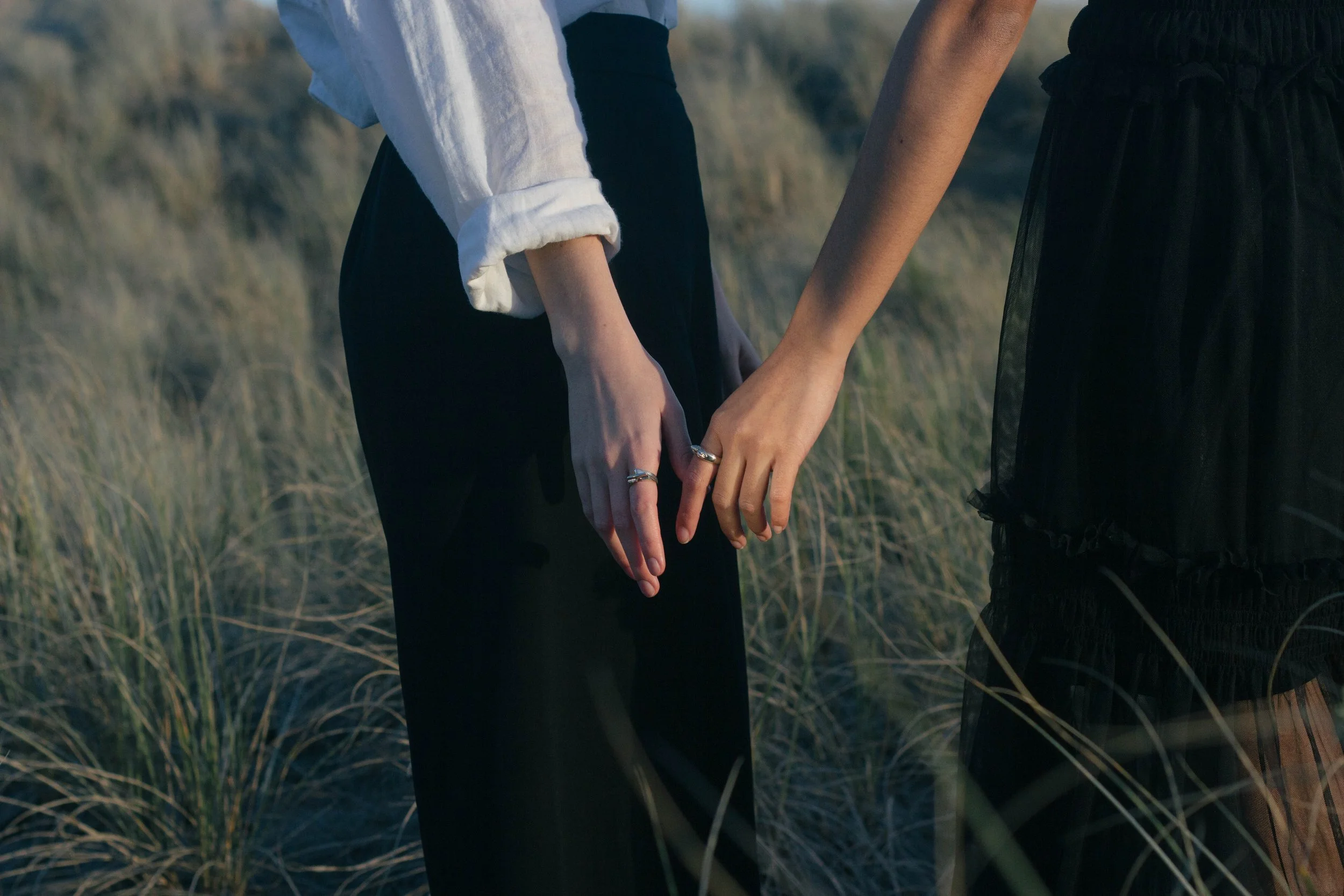 Two women wearing Mac Graham silver rings are holding hands at Bethels Beach Auckland, with only their hands, arms, and part of their outfits visible.
