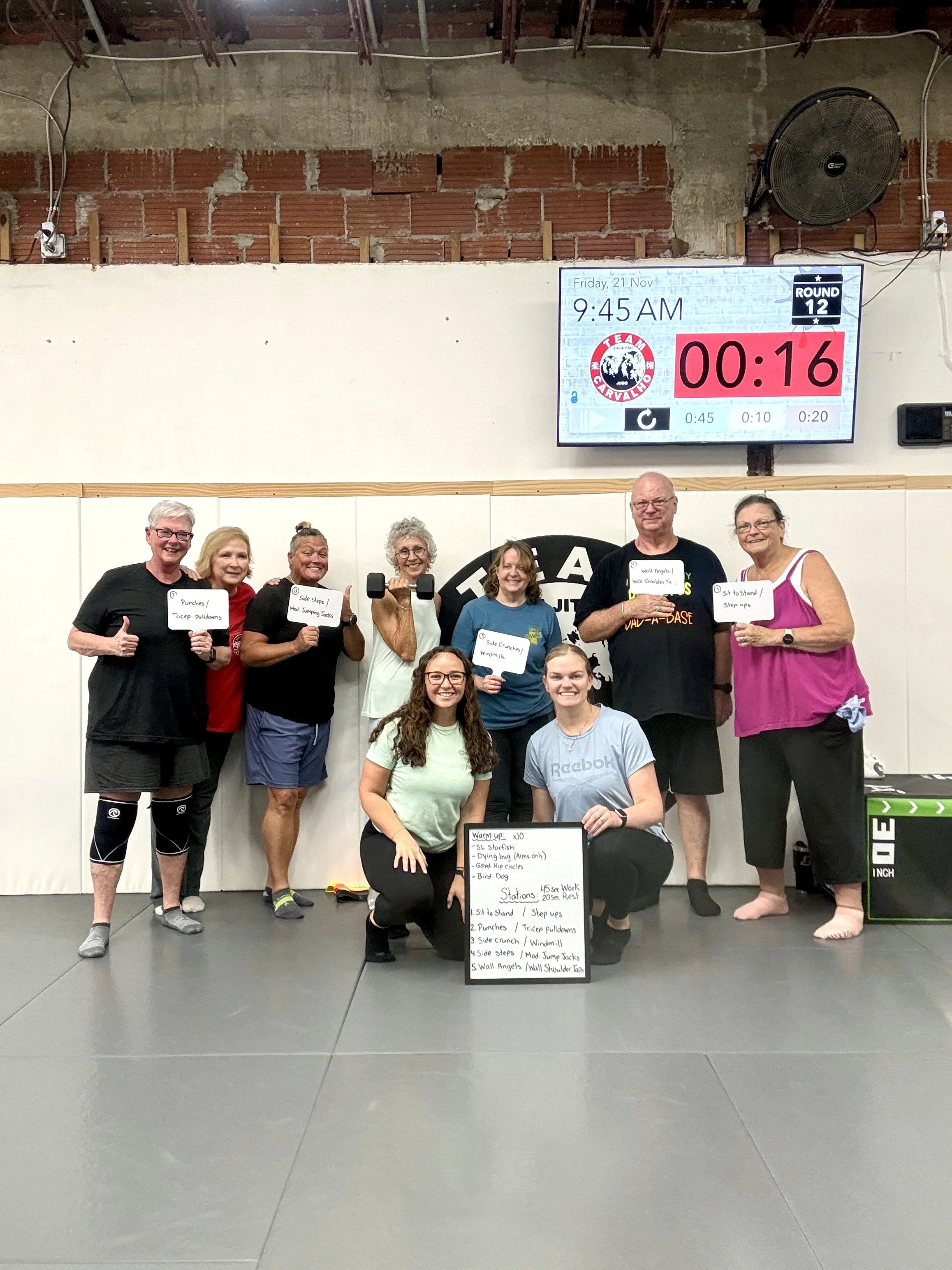 Group of people in a gym, posing with signs and workout equipment, celebrating after a workout session.