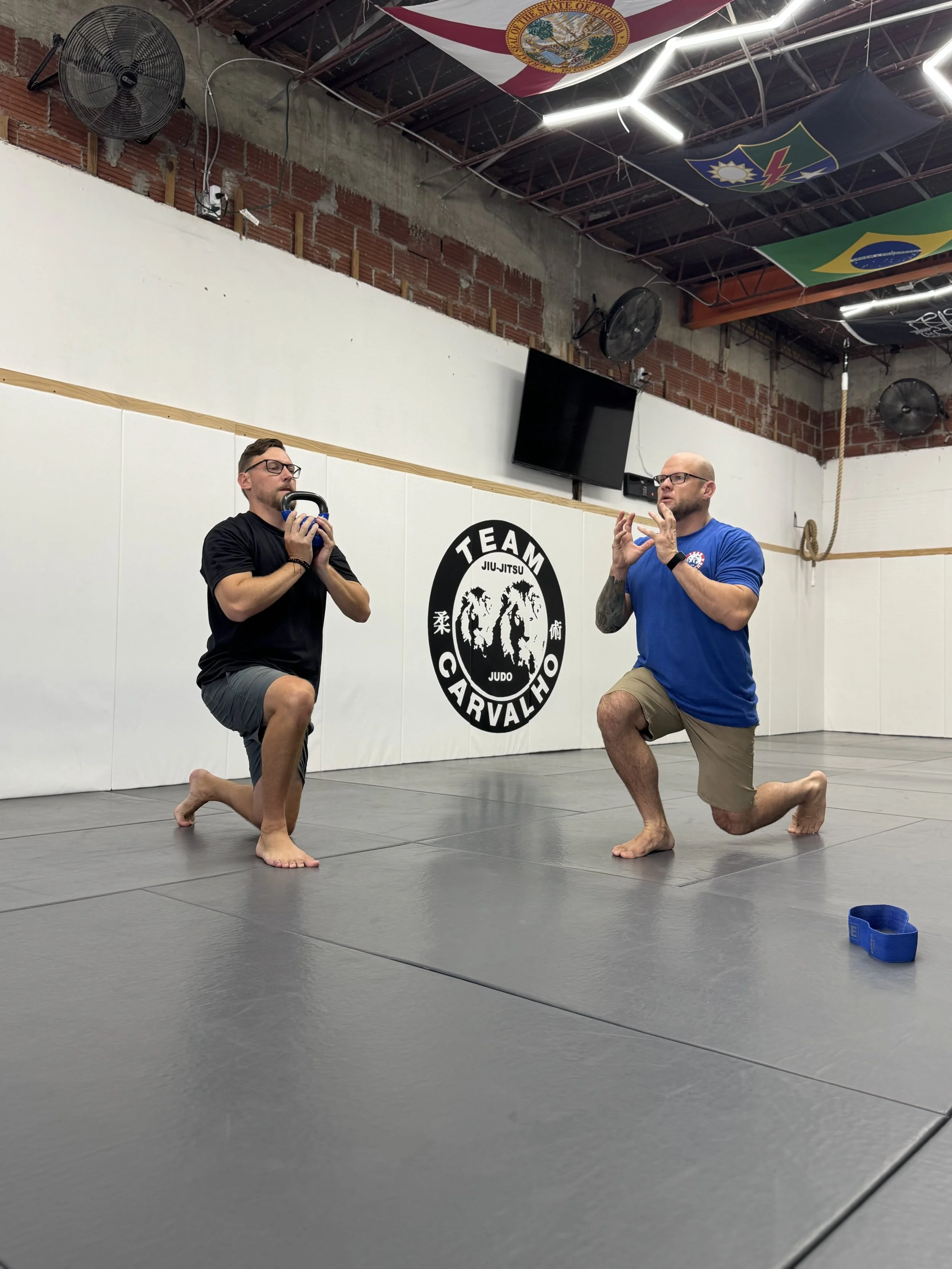 Two men are practicing martial arts in a gym with white walls and logos. One man is holding a kettlebell, and the other is speaking or instructing. They are both kneeling on one knee on the mat, and there are flags hanging from the ceiling.