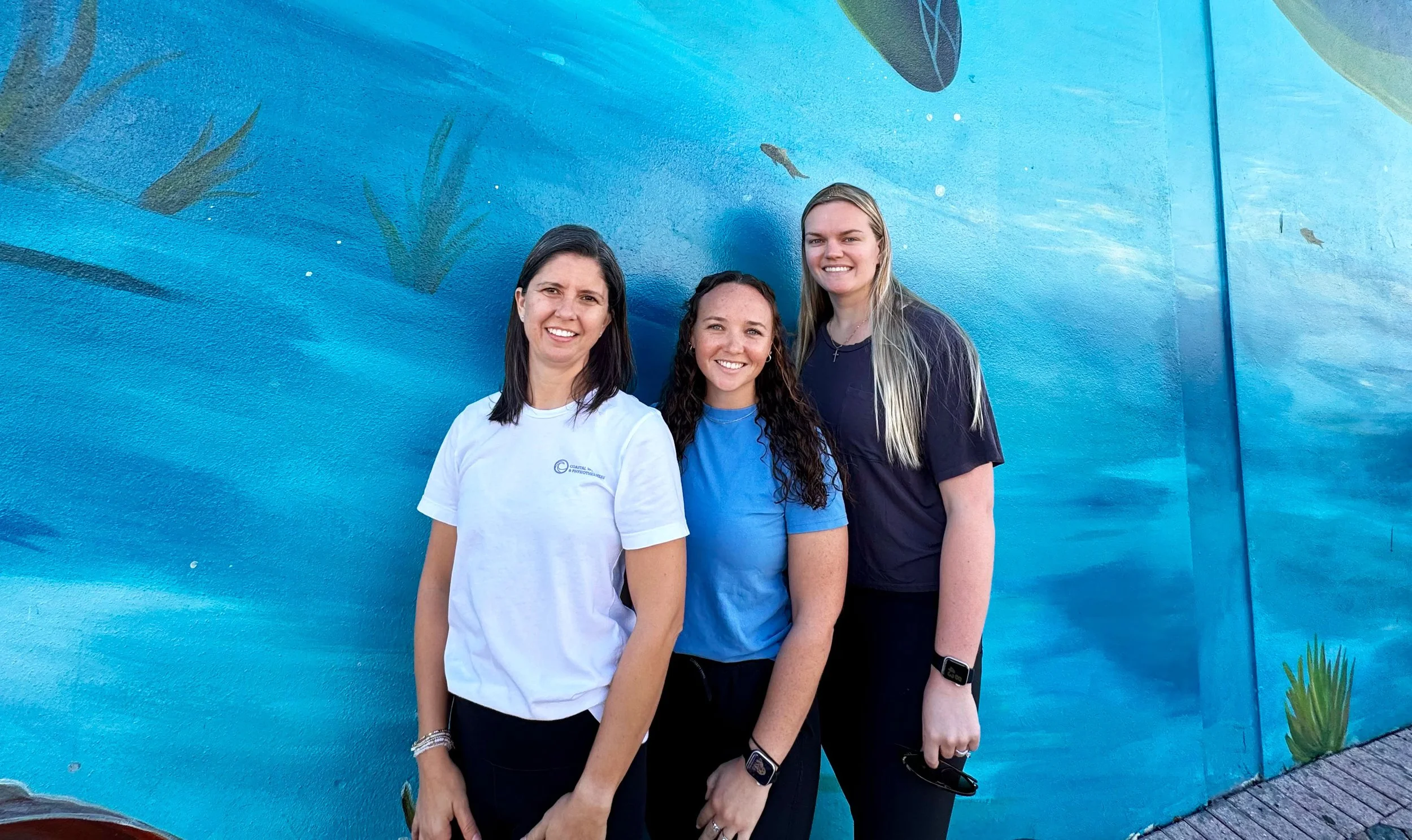 Three women standing in front of a blue mural wall with aquatic and plant imagery, smiling and posing for a photo.