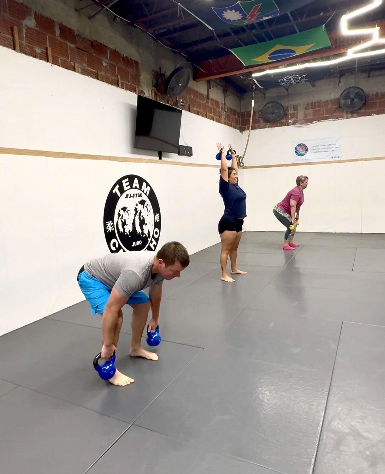 Three people exercising in a gym, with two holding kettlebells and one using a kettlebell while bending over, inside a martial arts studio.
