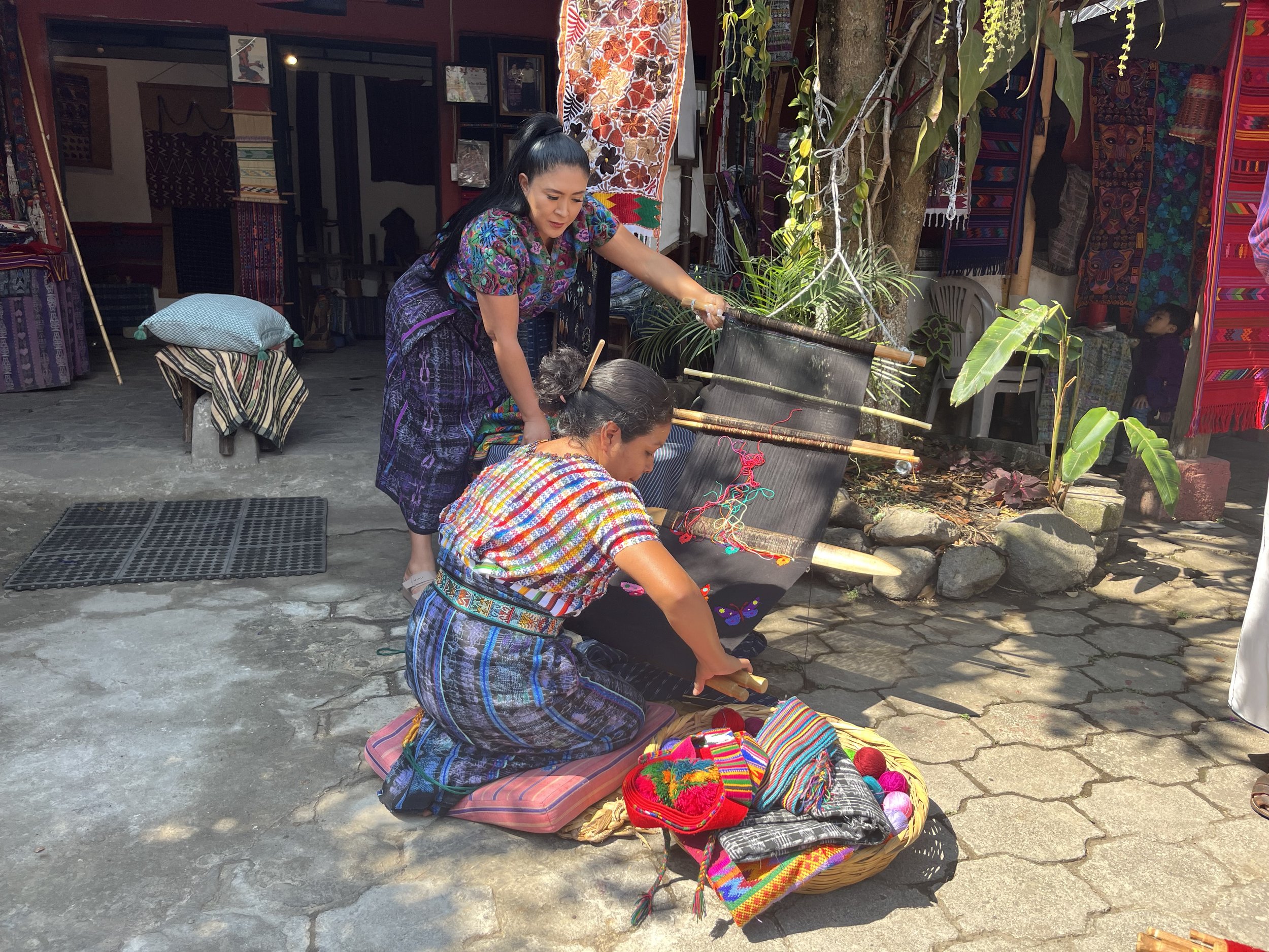 A woman weaving with a backstrap loom in Guatemala, wearing tradtional Guatemalan dress. Another woman holds the loom taught to show what she is doing.