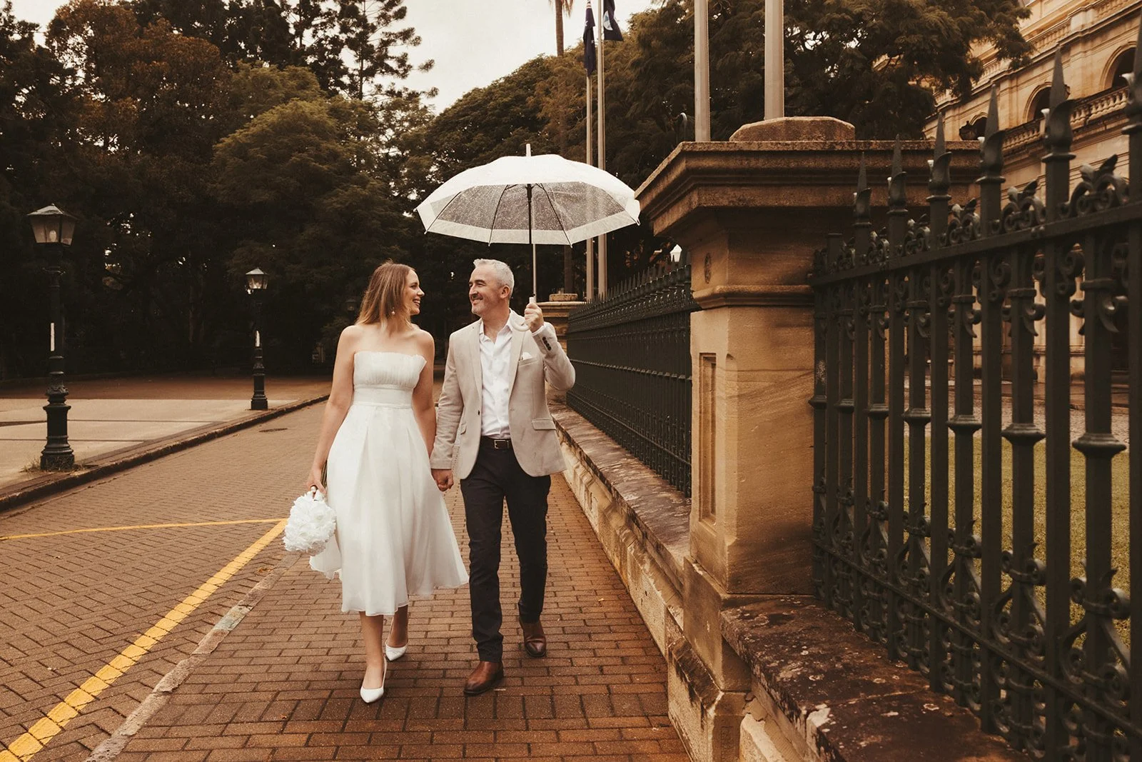 A couple, dressed in wedding attire, walks hand-in-hand on a brick sidewalk in a rainy outdoor setting, holding an umbrella and smiling at each other.
