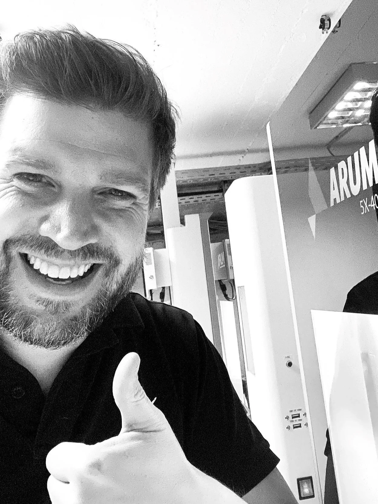 Ein lachender Mann mit dunklem Haar und Bart, der einen Daumen nach oben zeigt, im Hintergrund eine CNC Fräsmaschine mit elektronischen Geräten und einem Schild mit der Aufschrift 'ARUM' in einem Dentalen Fräszentrum.