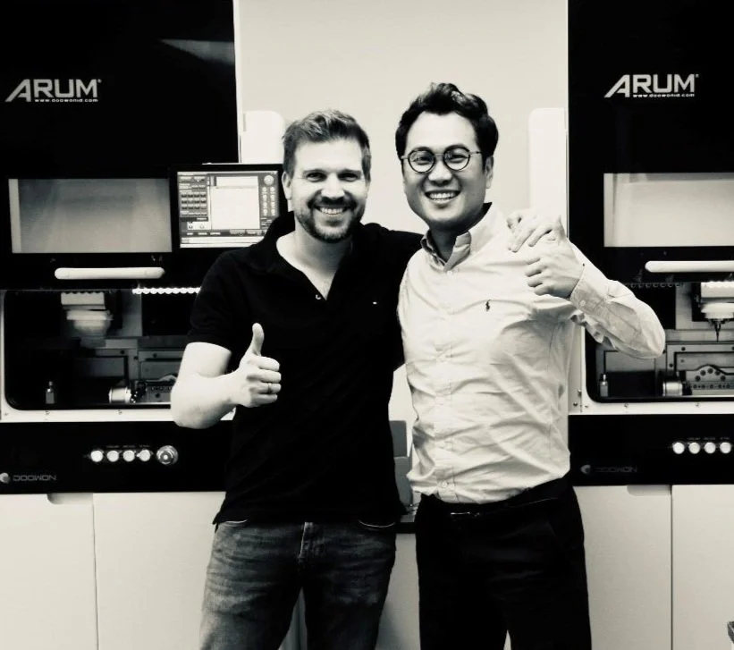 Zwei Männer posieren lächelnd vor Maschinen mit Daumen hoch. Hinter ihnen sind Geräte mit der Aufschrift "ARUM" sichtbar. Schwarz-weiß Foto.