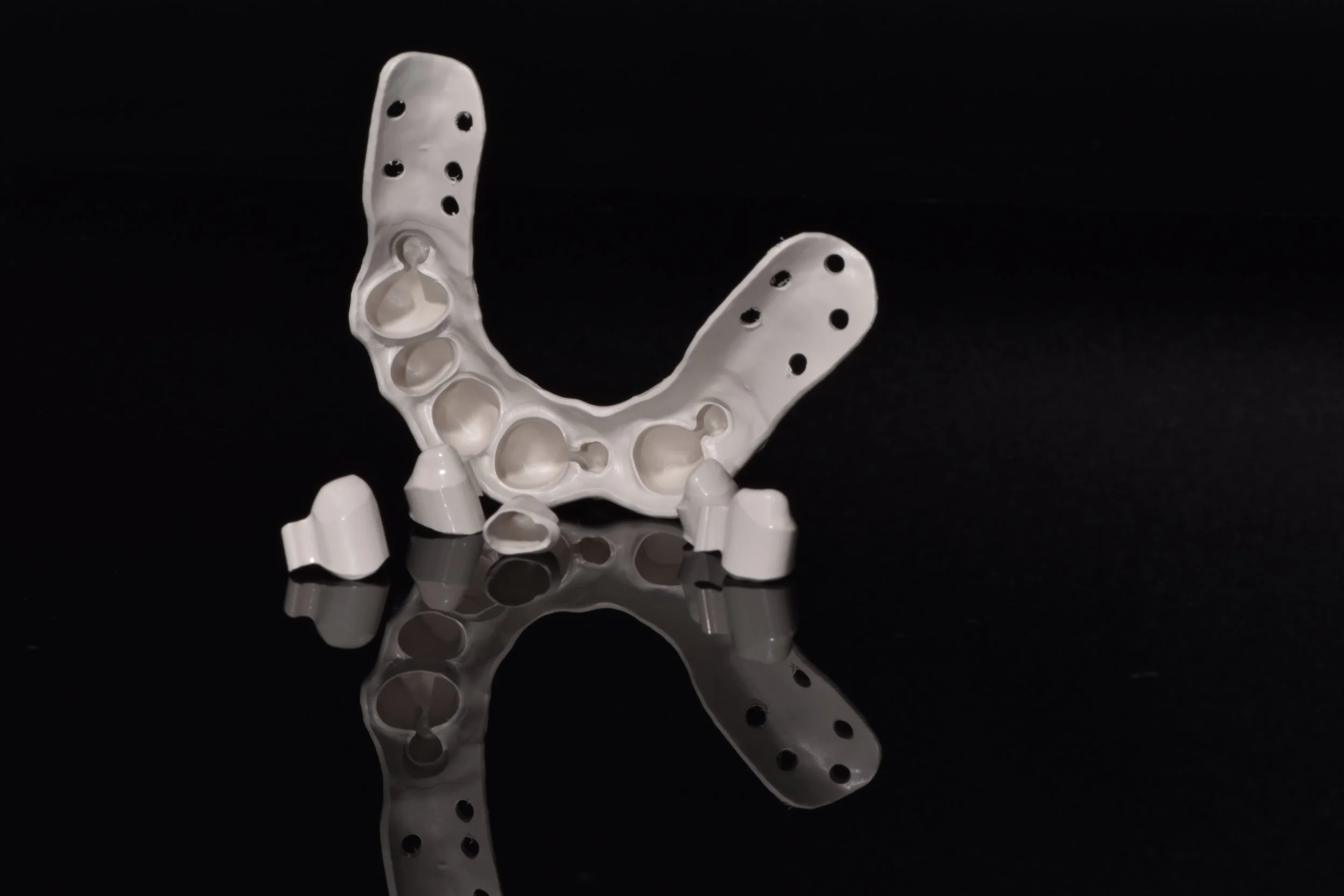Zahnersatz auf schwarzem Hintergrund, bestehend aus einem weißen Zahnimplantatgerüst mit Löchern und mehreren Attachments.