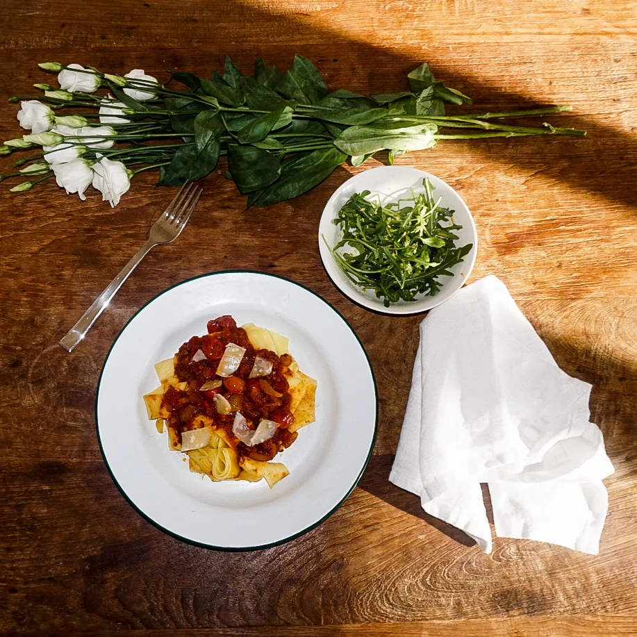 Rustic wooden table with loosely scattered white roses, a white plate with Tomato and Anchovy pasta, a white side bowl of rocket salad and white serviette