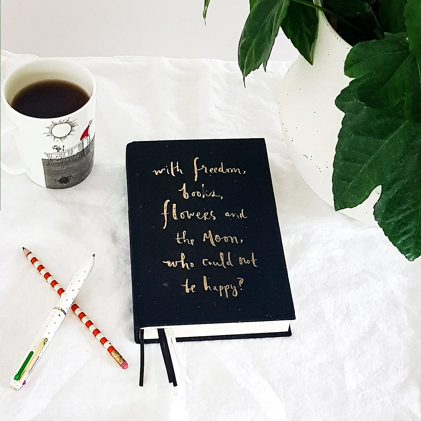 Flatlay to showJournal for shopping ban/ spending with pencil, pen beside it. Also mug with black coffee and pot plant. All on a white cotton table cloth. .