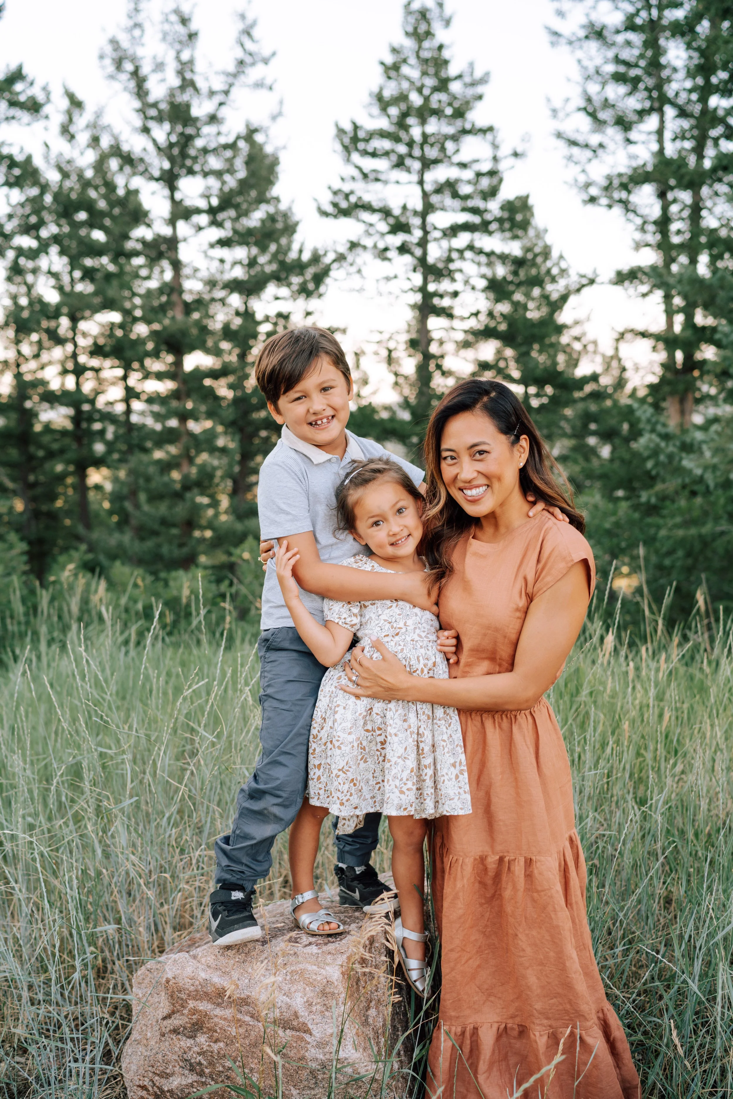 Mom in a rust-colored dress hugging her son and daughter among tall grass and pine trees during a Mount Falcon family session.