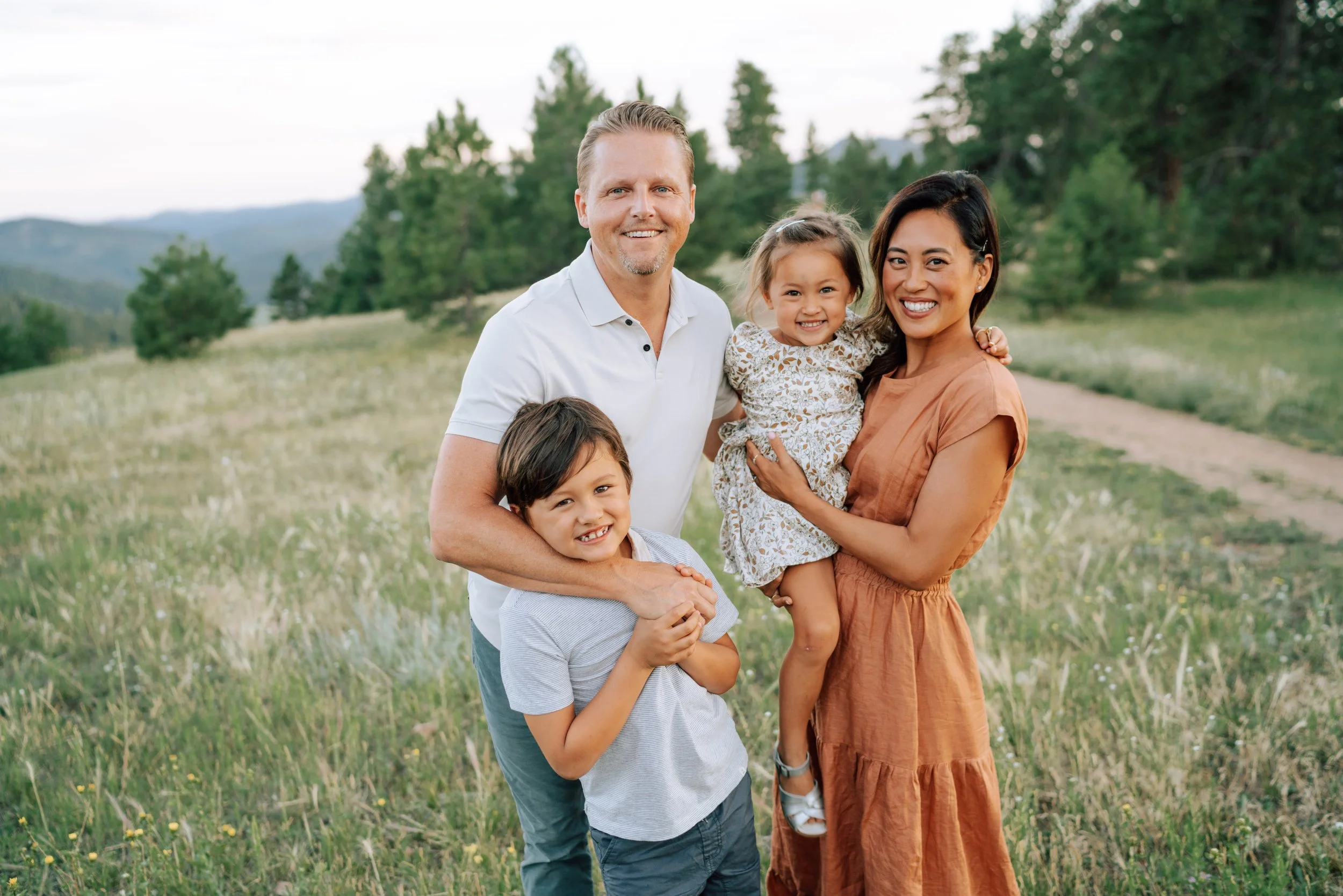 Family of four smiling together in a grassy mountain meadow with evergreen trees and Colorado foothill views.